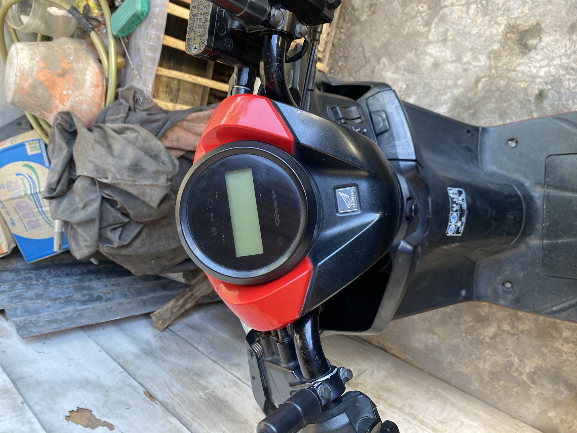 The image is a close-up, eye-level view of a black and red Honda scooter parked on a concrete ground. The scooter's dashboard is prominent, featuring a digital display screen, a circular speedometer, and the Honda logo. To the left of the dashboard are the scooter's handgrips and controls. In the background, a weathered wooden wall is visible, along with some scattered debris including a blue bag, a bucket, and a green brush. The overall impression is of a scooter parked in a somewhat cluttered outdoor area, possibly a backyard or workshop, in Bantaeng, Indonesia.