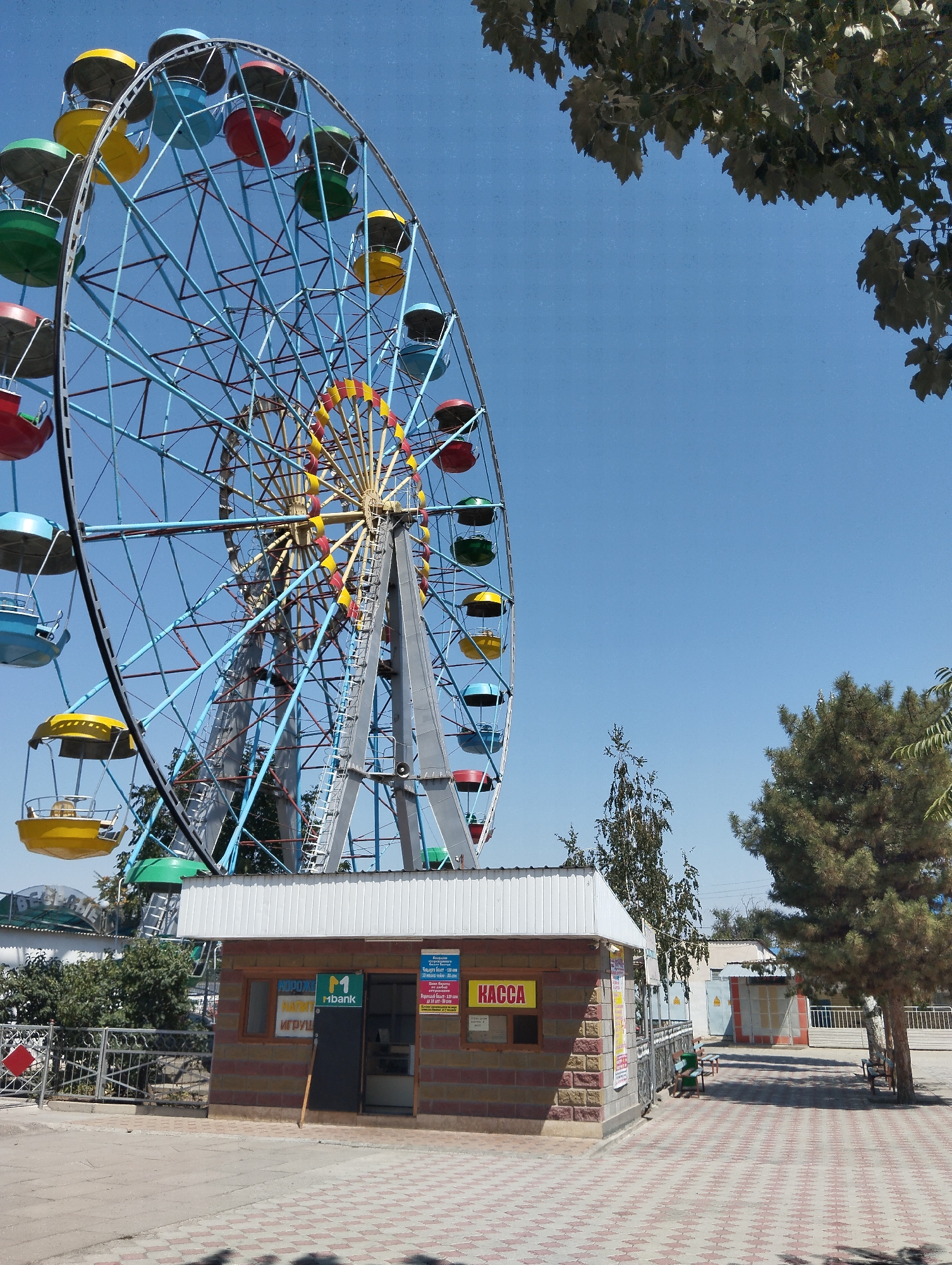 A Ferris wheel stands tall against a clear blue sky in an amusement park setting. The Ferris wheel is blue with brightly colored gondolas, including red, yellow, green, and blue. In the foreground, a small brick building with a white roof has signs in Russian, including one that says "KASSA" (which means "cashier" or "ticket office") and another with an "Mbank" logo. Adjacent to the building are patterned paving stones and some trees, one of which has large, green leaves that partially obscure the top of the Ferris wheel. There are no people visible in the image.