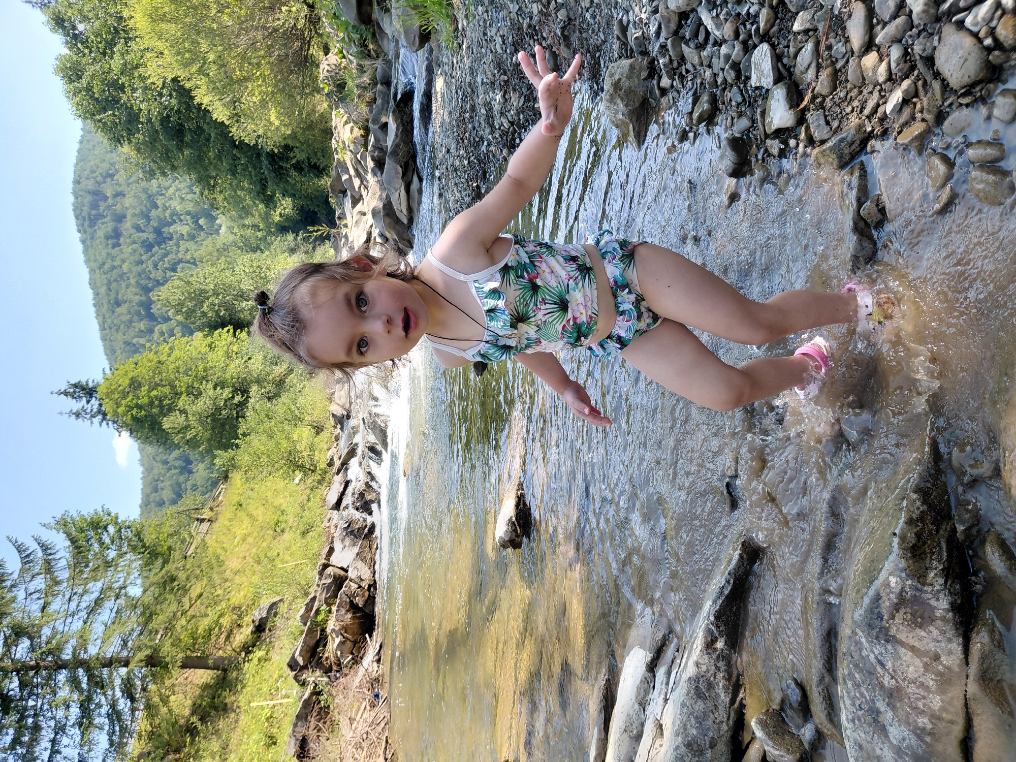 A young girl, approximately three to five years old, stands in a shallow, rocky river in a rural mountainous area. She is wearing a floral-patterned swimsuit and pink water shoes. Her expression is one of surprise or excitement. Her hands are outstretched. The river flows over smooth stones and smaller rocks. The background features lush green trees and a forested mountain slope under a clear blue sky. The location is identified as Chernyk, Ukraine.