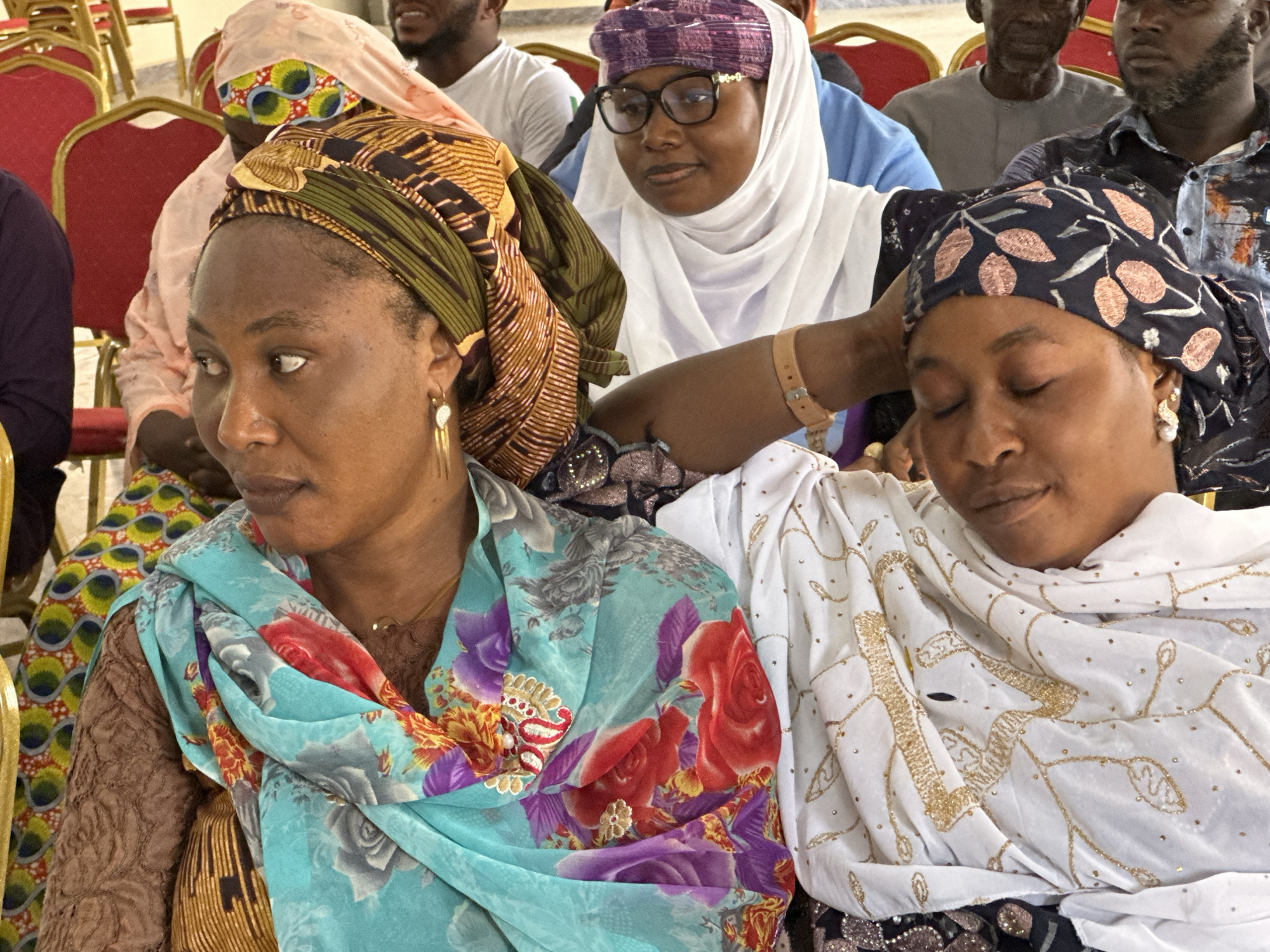 The image shows three women in an indoor setting, likely a meeting or gathering based on the presence of chairs and other people in the background. The woman on the left wears a colorful headwrap and a turquoise scarf with floral patterns, while the woman on the right wears a patterned headwrap and a white shawl with gold embroidery. The woman in the background wears a purple head covering, eyeglasses, and a white hijab. The location is Unknown, Unknown.
