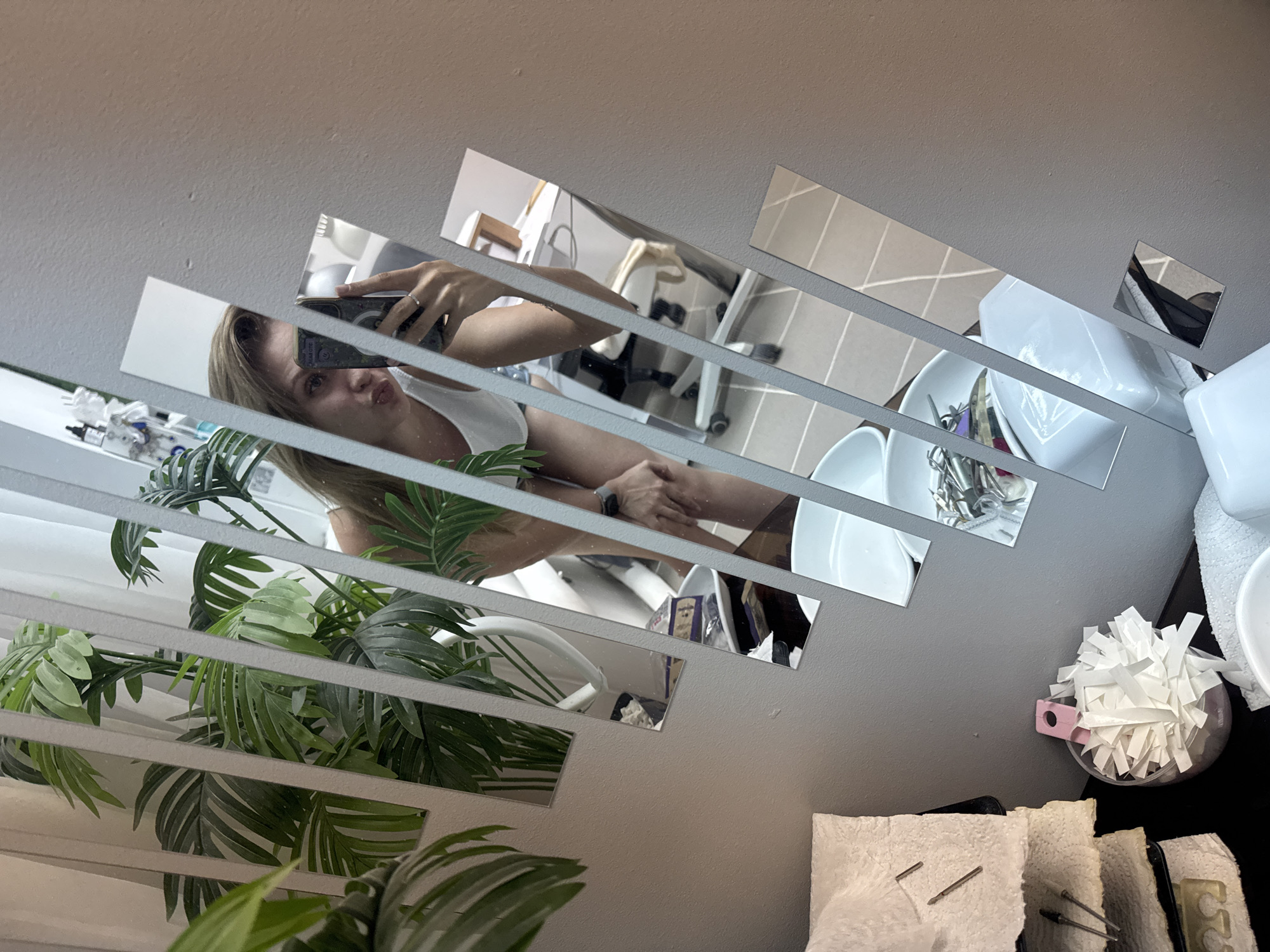 A woman is taking a selfie in a segmented mirror, likely in a bathroom. The mirror reflects a scene including a sink with various containers and items, possibly related to cosmetics or personal care. A green leafy plant is partially visible on the left side of the mirror. The mirror is mounted on a grey wall. Based on the reflection, the room appears to be a typical bathroom setting. There is no indication of the city of Izhevsk, Russia, in the image.