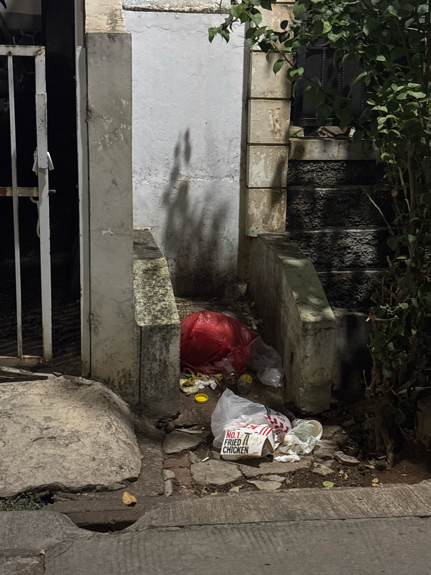 The image depicts a street scene at night in Bekasi, Indonesia, characterized by a collection of trash. In the foreground, a grey concrete surface with a drainage channel is visible. To the right, scattered trash includes a red plastic bag, plastic bottles, a crumpled white plastic bag, and a white bucket with the "NO.1 FRIED CHICKEN" logo, likely from KFC, along with other unidentifiable debris. A beige concrete pillar stands to the left, adjacent to a white metal gate. Behind the pillar and the trash, a set of rough, dark stone stairs ascends, partially obscured by dense green foliage on the right side of the image. The overall environment appears neglected and littered.