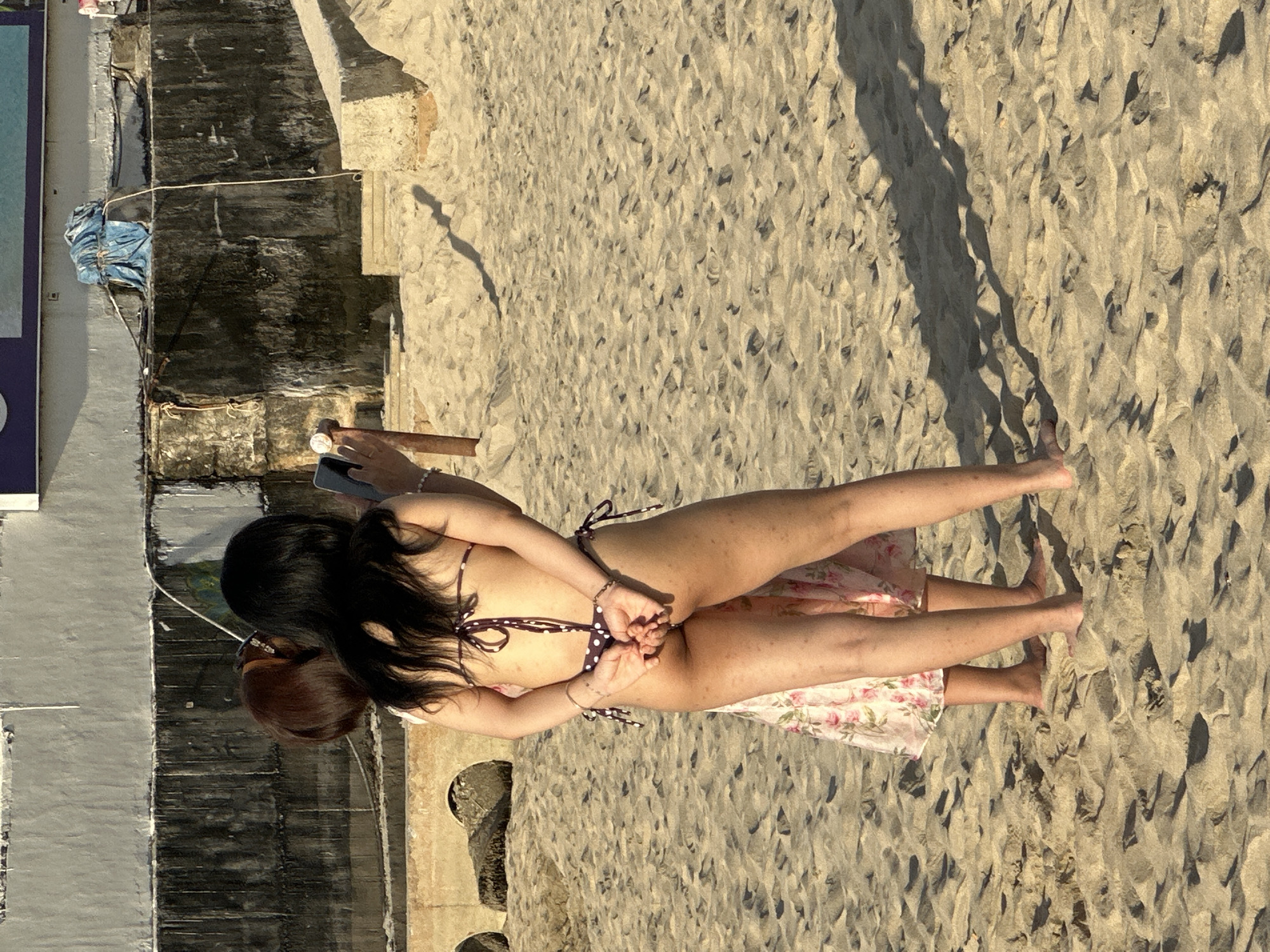Two individuals are on a sandy beach in Da Nang, Vietnam. One person, seen from the rear, wears a dark polka dot bikini and has their hands clasped behind their back. They are holding a smartphone in their right hand, appearing to take a picture or record a video. The other person, positioned to the left and partially obscured, wears a light-colored floral dress. The background features a concrete wall with some graffiti and water damage, and further back, a building with a blue tarp secured by ropes. The sand is uneven with visible footprints and shadows cast by the individuals and possibly nearby structures. The lighting suggests it is daytime.