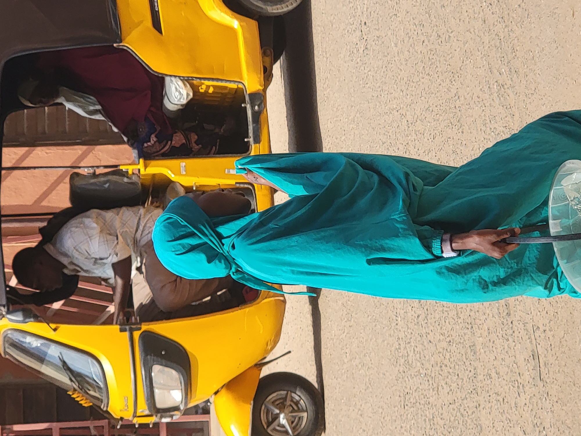 In Maiburiji, Nigeria, a yellow Bajaj Qute auto-rickshaw is parked on a paved surface. Two passengers are inside, one in the driver's seat wearing a light-colored, collared shirt. To the side of the vehicle, a woman wearing a teal headscarf and matching garment stands on the pavement. She is holding a long, dark object, and a partially visible, transparent container.