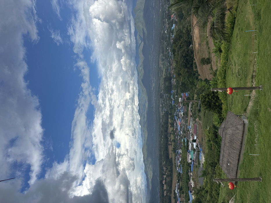 The image presents a panoramic landscape view of Ban Mai Saha Sampan, Thailand, captured from an elevated position under a dynamic sky. The upper approximately 60% of the frame features a bright blue sky partially covered by large, distinct cumulus clouds, varying in color from white to dense gray. A range of tree-covered mountains forms the horizon line in the background. Below the mountain range, the midground displays a valley containing the settlement of Ban Mai Saha Sampan, characterized by numerous residential structures with varied colored roofs, including blue, red, and gray, interspersed with green trees and cultivated fields. The foreground consists of a sloping, manicured grassy area. Centrally located in the lower foreground is a small, open-sided pavilion or hut with a distinctive multi-tiered, thatched or tiled roof. Two vertical wooden posts are present in the immediate foreground, positioned to the left and right of the hut, each supporting a spherical red lantern. A segment of a palm frond is visible on the far right edge of the frame. No individuals, actions, or specific interactions are observed within the scene.
