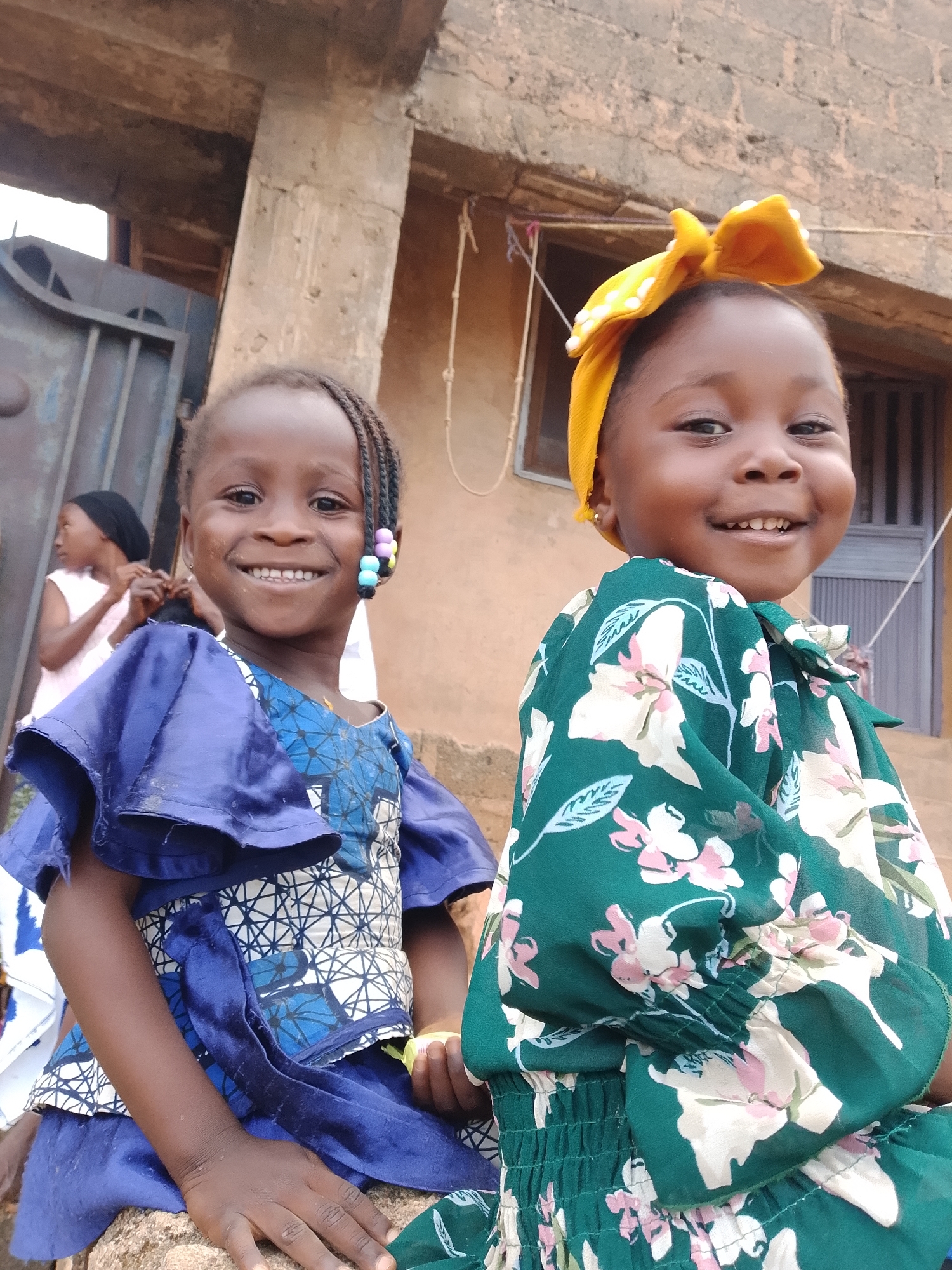 Two young girls are sitting outdoors in front of a building with a weathered, concrete exterior. The girl on the left has braided hair adorned with colorful beads and is wearing a blue dress with intricate patterns. She is smiling broadly, showing her teeth. The girl on the right is wearing a yellow bow on her head and a green floral-patterned dress. She is also smiling. In the background, another person, a young girl wearing a black headscarf and a white dress, is visible. The environment suggests a casual, possibly residential setting in a town or city.