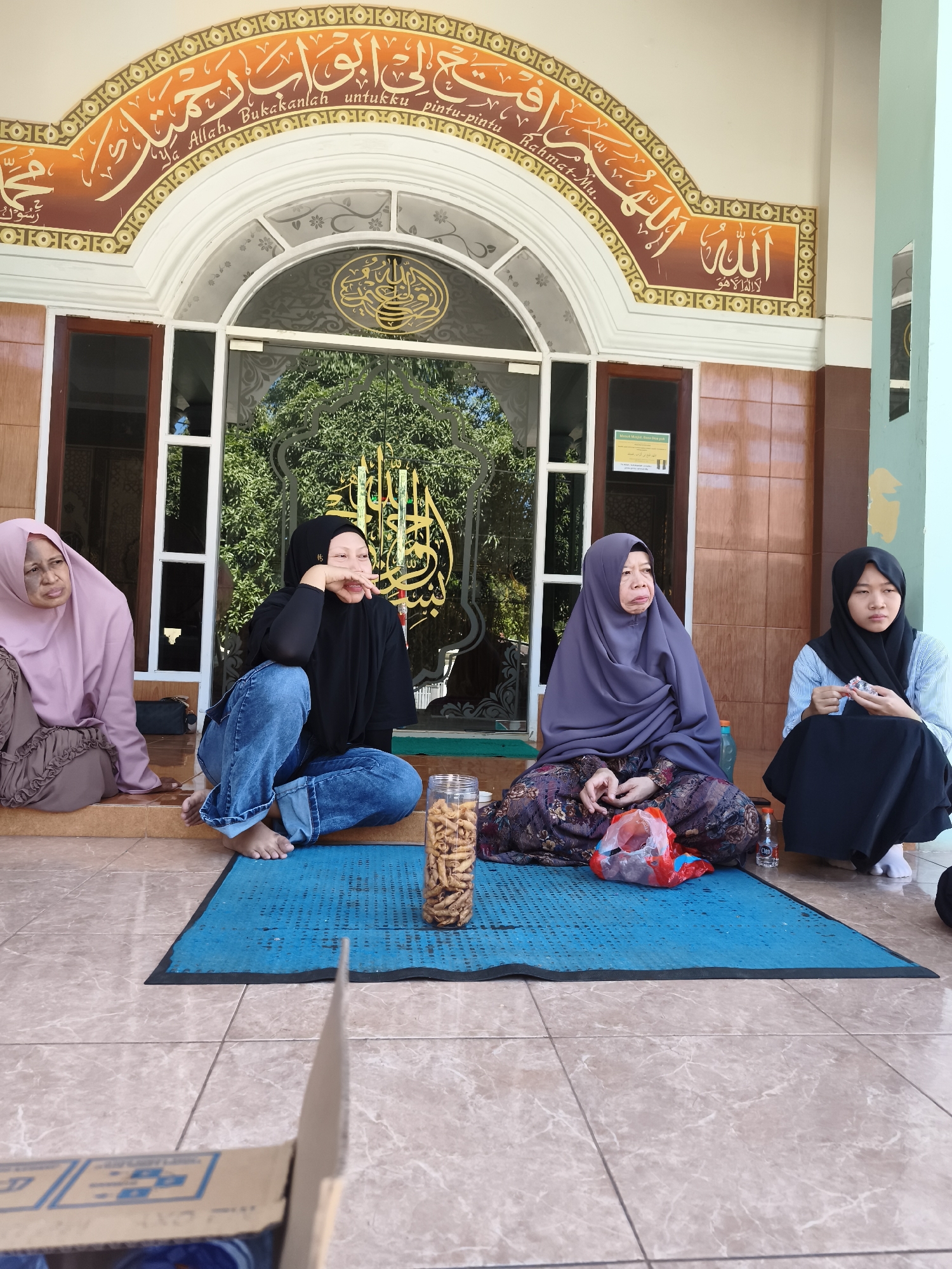 Four women are seated on a blue mat outside a building with arched doorways and windows. The women are all wearing headscarves, with three of them wearing full-length dresses and one wearing jeans and a t-shirt. One woman on the far left is wearing a light pink headscarf and a ruffled brown dress, looking to her left with a concerned expression. Next to her, a woman in a black headscarf and blue jeans is looking to her right with her hand near her mouth, seemingly in thought. The third woman, in the center, is wearing a dark purple headscarf and a patterned dress, looking forward with a neutral expression. The woman on the far right is wearing a black headscarf and a light blue and white striped shirt, also in a black skirt, holding something small in her hands. In front of the women are a jar of snacks, a red plastic bag, and a bottle of water. The building behind them features ornate Arabic calligraphy above the entrance, suggesting a religious or cultural site, likely a mosque, given the context of Tonasa, Indonesia, a region with a significant Muslim population. The flooring appears to be tiled, and there is greenery visible through the glass doors.