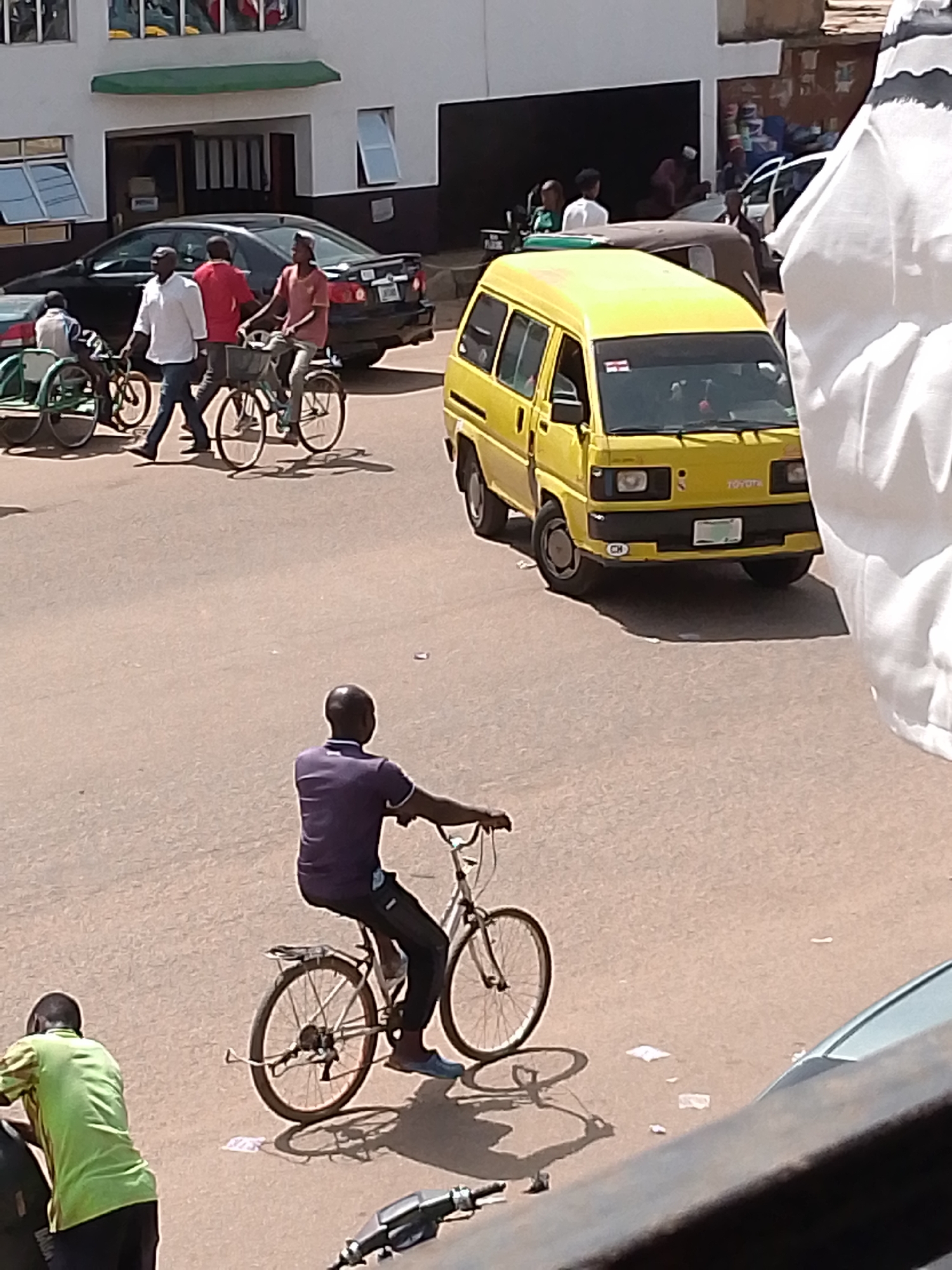 In Bukuru, Nigeria, a busy street scene unfolds under what appears to be a bright, sunny day. The foreground is dominated by a man riding a bicycle, dressed in a purple polo shirt and dark pants. His bicycle is a typical road bike with thin tires and a rack over the rear wheel. To his left and slightly further back, another individual in a bright green shirt is bent over something that is partially visible, possibly a motorcycle.
Further down the street, several other individuals are engaged with bicycles. Two men are walking beside a bicycle that has a basket attached to its handlebars. One of them wears a white shirt and dark pants, while the other is in a red shirt and lighter pants. Behind them, another person in a maroon shirt is also on a bicycle, moving in the same direction. These individuals and their bicycles are in front of a dark sedan, which is parked or moving slowly. In the background, a yellow minibus, possibly a Toyota, is also visible, suggesting public transportation is in operation. The scene appears to be in an urban or semi-urban environment, with buildings and paved roads. The sunlight casts distinct shadows, reinforcing the impression of a clear day. The overall atmosphere is one of everyday activity and movement.