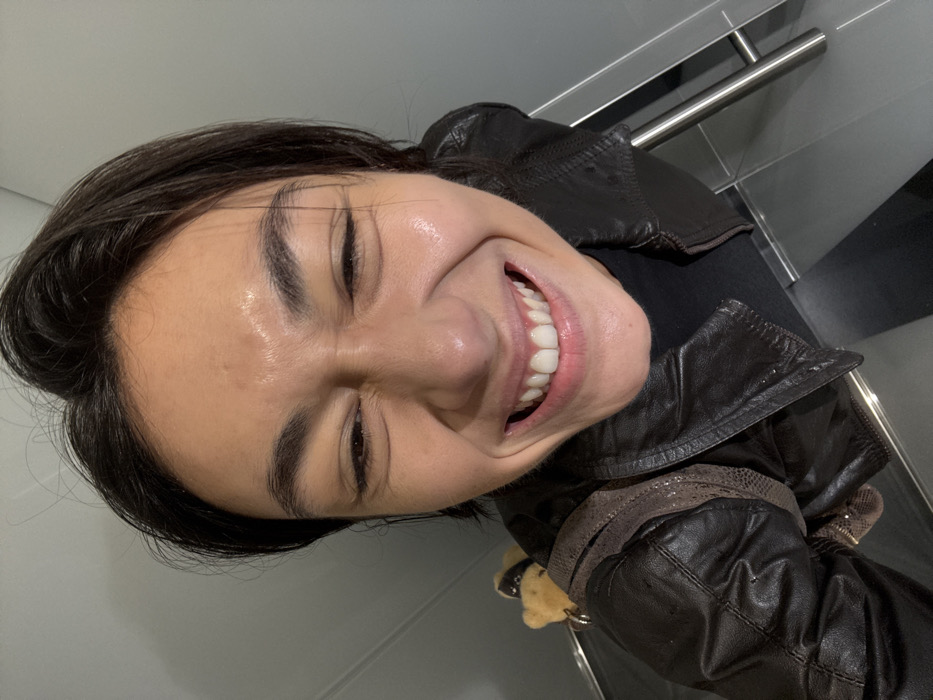 A close-up, low-angle selfie depicts a single individual, appearing to be an adult female, from the upper chest upwards. Her face is centered, exhibiting a wide, joyful expression with tightly squinted eyes, prominent cheek lines, and an open mouth revealing a full set of white teeth. Her skin has a visible sheen. She wears a dark brown leather jacket over a black garment. A textured, dark-patterned strap, possibly from a bag or backpack, is visible over her left shoulder, to which a small, light brown teddy bear charm adorned with a dark harness or collar is attached. The background is minimalist, consisting of plain light gray walls and a dark, reflective surface below. A silver metallic handle is partially visible on the right side of the frame. The specific location within Paris, France, cannot be determined from the visual evidence provided.