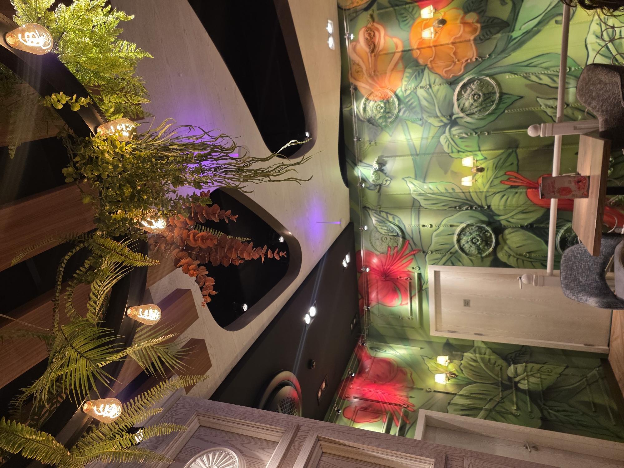 The image shows the interior of a restaurant or cafe. The view is from a low angle, looking upwards towards the ceiling and walls.
On the left, there's a decorative arrangement of greenery, ferns, and hanging lights. The light fixtures are modern-style bulbs that are exposed, and the greenery is dense. The decor is mounted on a geometric structure, possibly part of the wall design.
On the right, the wall is covered with a mural depicting flowers, leaves, and other botanical elements. The mural has a whimsical, almost cartoonish style. There is a door in the middle-right.
The space appears to be designed with a combination of natural and artificial elements, creating an eclectic and atmospheric ambiance.
Without additional information, the location cannot be pinpointed in Plovdiv, Bulgaria.