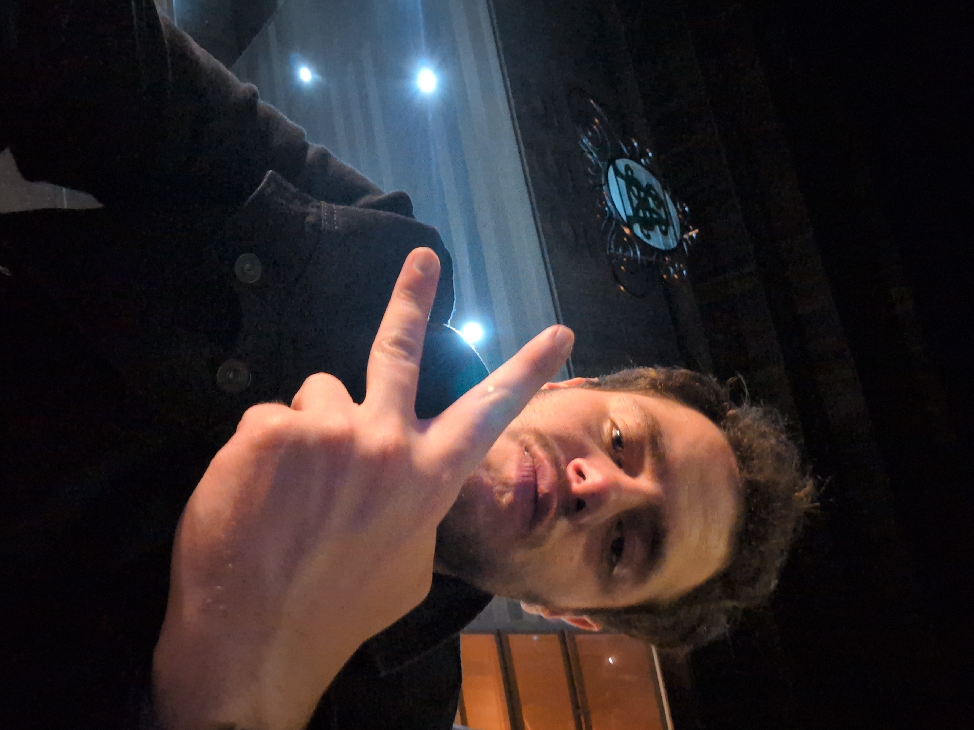 A selfie of a man is positioned with his face toward the camera, slightly turned, his right hand raised making a peace sign. He has curly dark hair and stubble on his face. He is wearing a dark button-down jacket. Behind the man, the background consists of reflective surfaces illuminated by lights and a decorative emblem in the upper-right corner. The location is Mexico City, Mexico.