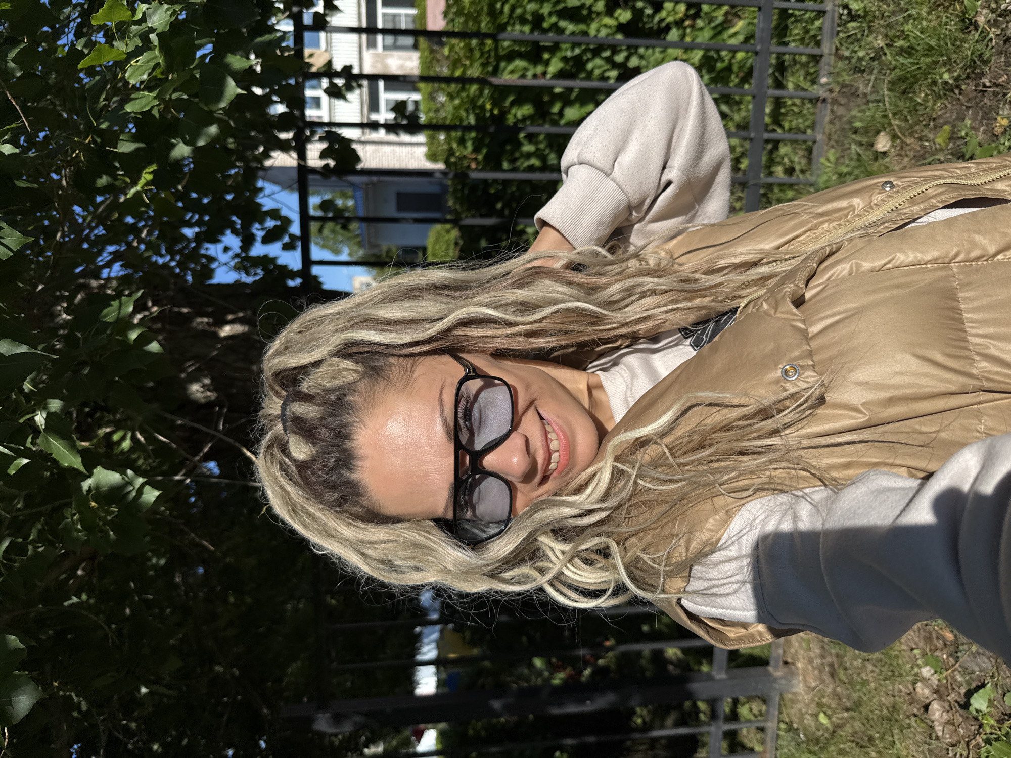 In Rusanovo, Russia, a woman with blonde, curly hair pulled into a high ponytail, wears dark-rimmed sunglasses, a beige vest over a light-colored sweatshirt, and is photographed outdoors. She is smiling and touching her neck with her right hand. A black metal fence is visible behind her, with green foliage and trees in the background. Sunlight casts a shadow on her left arm.