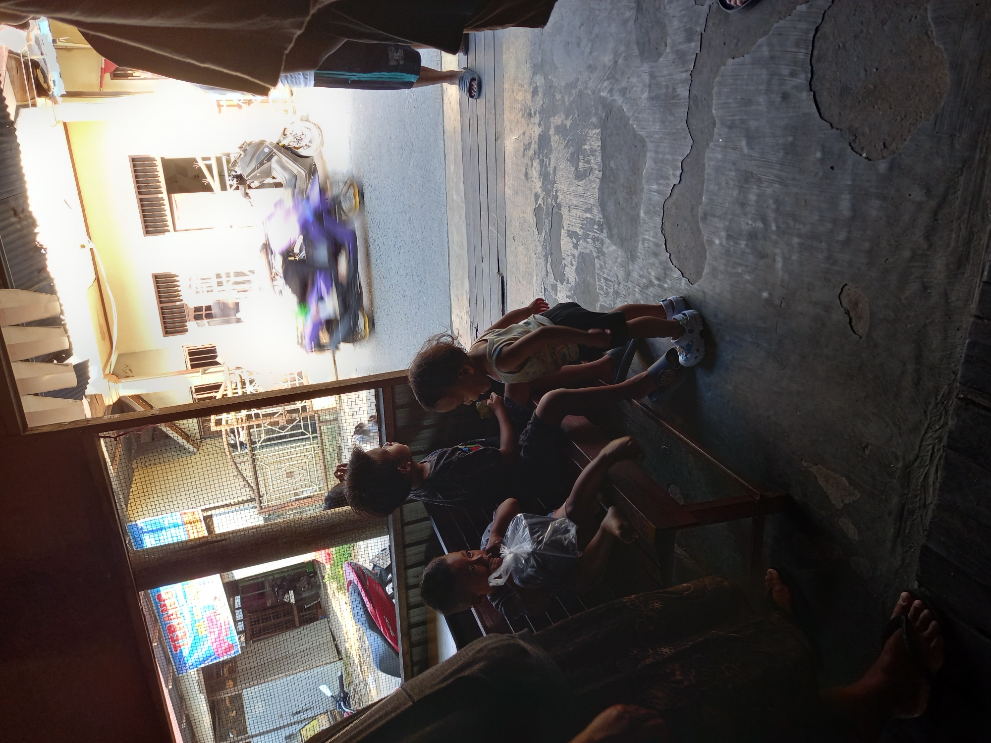 A medium shot captures an outdoor scene in Samarinda, Indonesia, likely a roadside establishment or a residential porch, given the presence of a wooden bench and a concrete floor with some peeling paint. In the foreground, a young child, wearing a light-colored tank top and dark shorts, stands on the concrete, looking towards the left. To the left of this child, on a wooden bench, sits another child holding a clear plastic bag filled with dark items. Further to the left, an adult's legs are visible, clad in dark fabric and sandals. Behind them, an older child sits on the bench, facing away from the camera and looking towards the right. The background reveals a street with a purple scooter in motion, creating a blur of color. Buildings line the street, and a red and white banner is visible hanging on one of them. The overall lighting suggests it is daytime, with strong sunlight casting shadows.