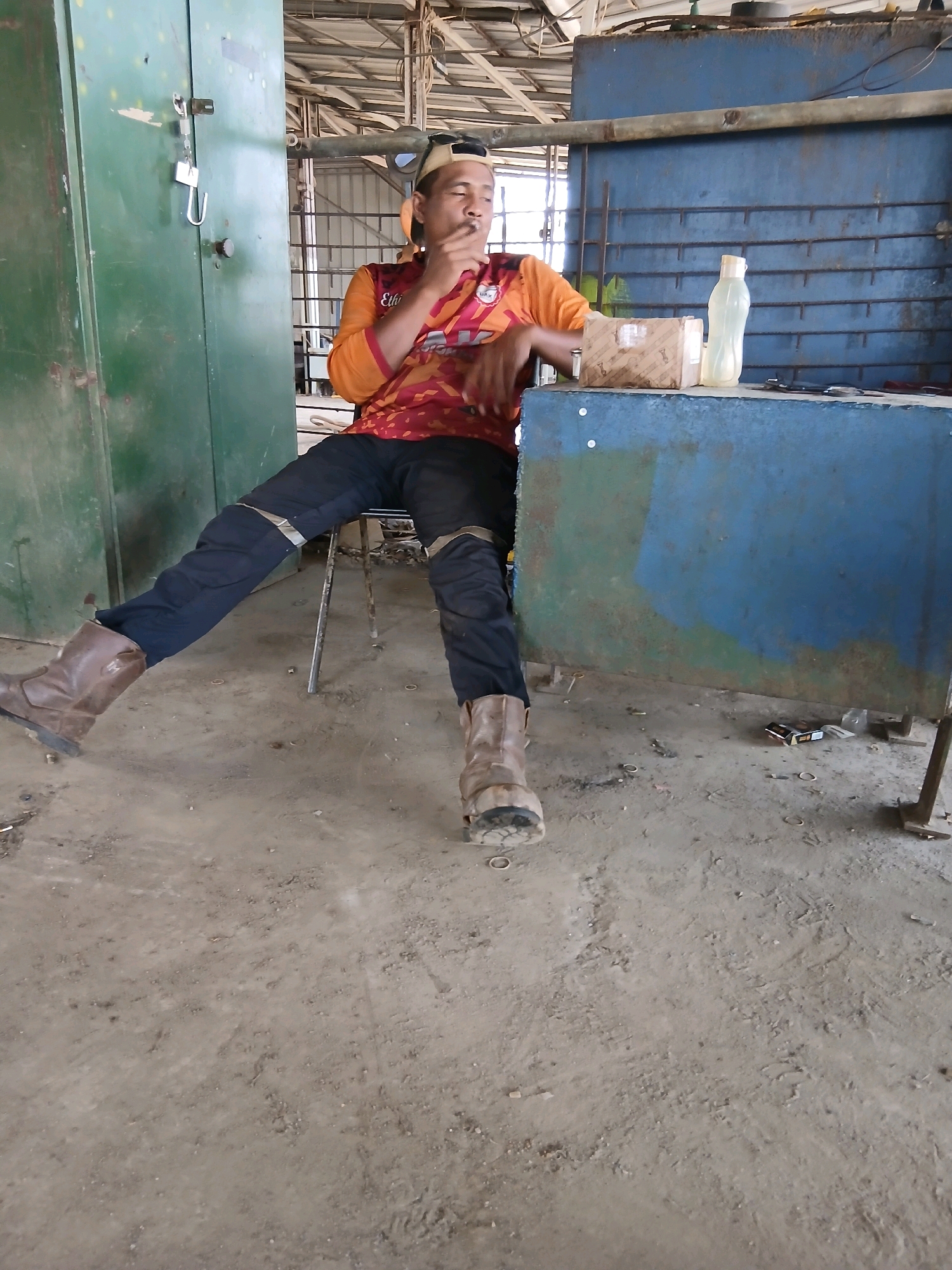 A man wearing a red and orange long-sleeved shirt, black pants, and brown boots sits on a metal chair. He is smoking a cigarette and looking down. He is seated at a blue metal desk with a cardboard box and a plastic bottle on it. Behind him, a large green metal cabinet is visible. The scene appears to be in an industrial or workshop setting, with a concrete floor and a metal ceiling structure. The overall lighting suggests an indoor environment with natural light sources. The location is identified as Pangkalpinang, Indonesia.