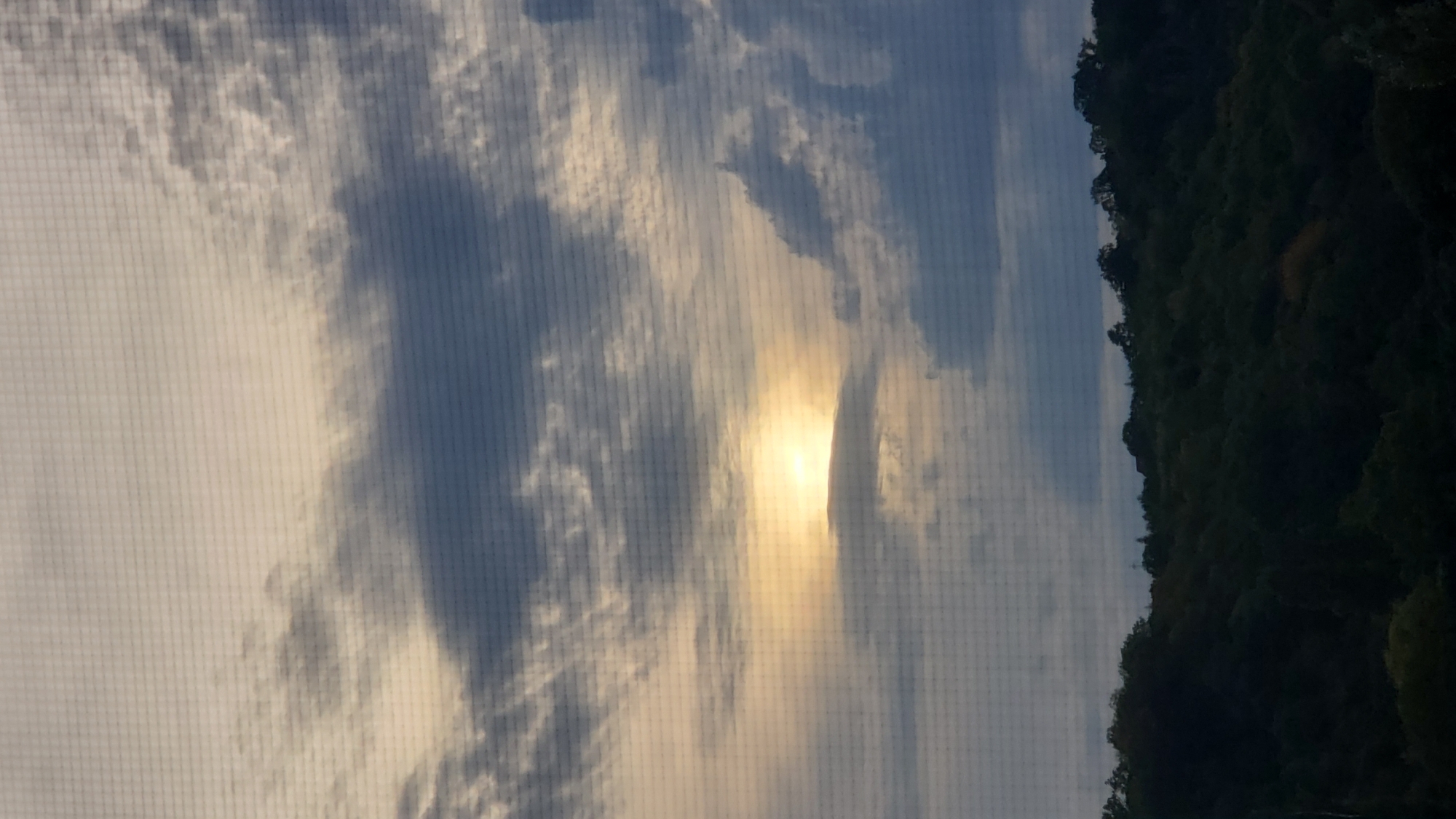 The image is a photograph of a cloudy sky over a dark, tree-covered hillside. The sun is partially visible through a break in the clouds, casting a warm, yellow glow. The sky is a mix of dark grey and lighter grey clouds, with streaks of white. The hillside is densely covered in trees, appearing almost black in the low light. A subtle grid pattern is visible over the entire image, suggesting it may have been taken through a screen. There are no people or specific structures visible in the image. The scene appears to be a natural landscape.