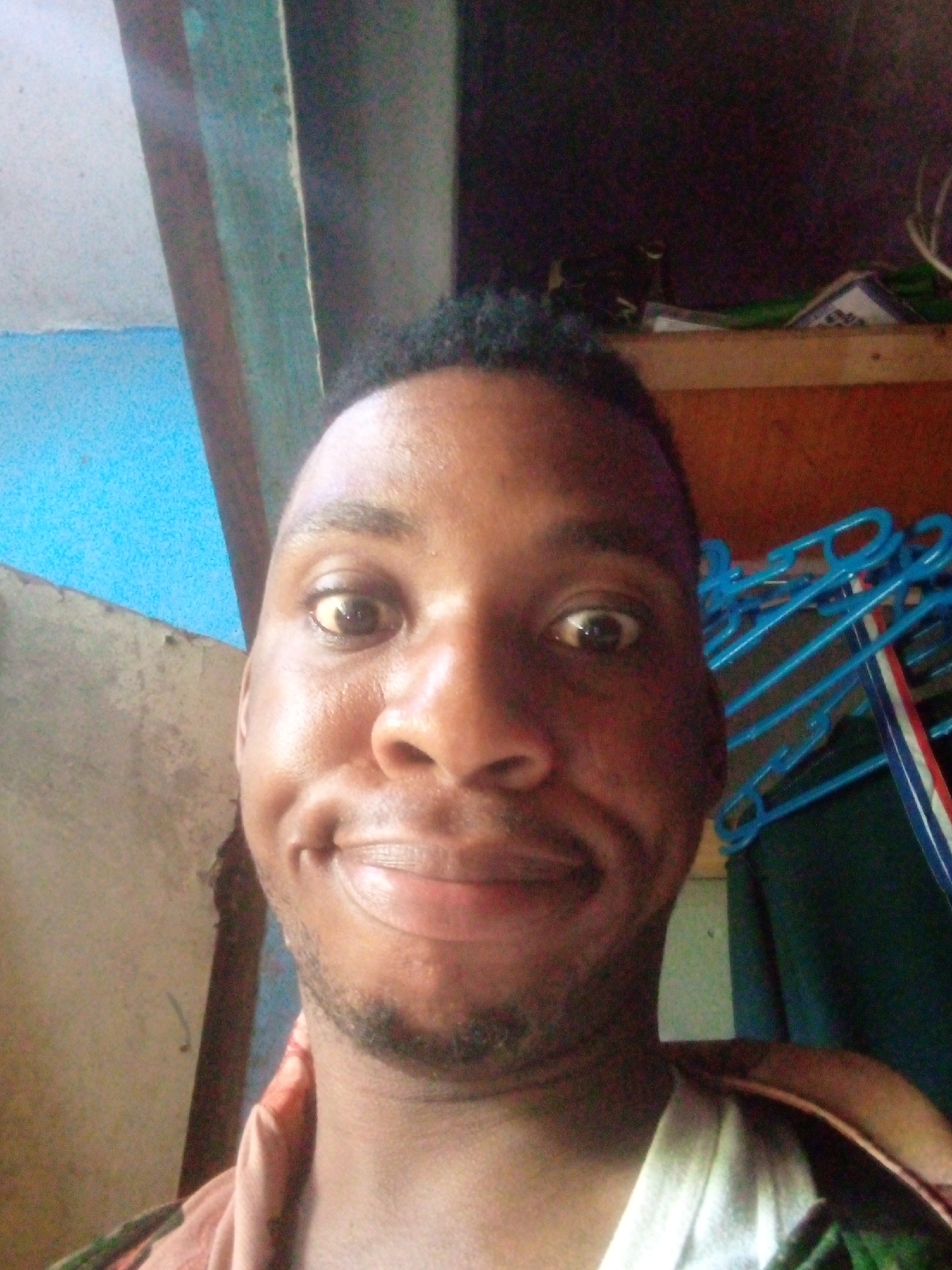 A close-up selfie photograph shows a smiling young African man indoors. The location is likely inside a room in Ikorodu, Nigeria, given the inferred prompt. The man has short black hair and a close-cropped beard. His skin appears slightly oily. He is wearing a t-shirt and a patterned shirt over it. In the background, there are clothes hangers, possibly hanging garments, a wooden shelf and walls. The lighting appears natural but limited. The paint on the wall next to the window is peeling off.