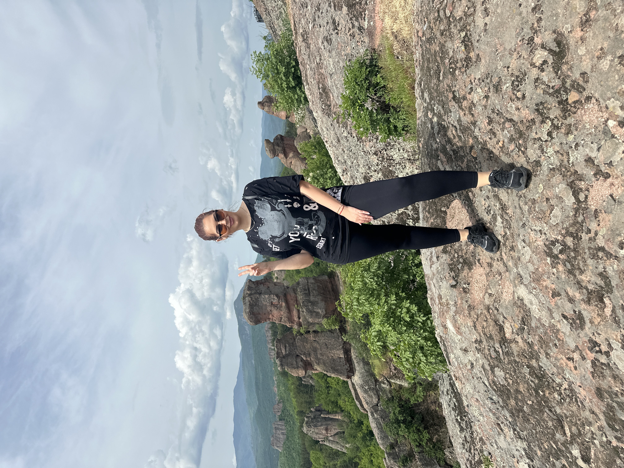 The image shows a woman standing on a rock formation, making a peace sign with her left hand. She is wearing sunglasses, a black t-shirt, and black leggings. Her right hand is positioned by her right hip. The woman is likely in the city of Belogradchik, Bulgaria, which is visible in the background and famous for its rock formations. The background features a mountainous landscape with distinct rock formations covered in vegetation. The sky is partly cloudy. The rock formation the woman stands on also has green vegetation and a rough texture. She is wearing black running shoes.