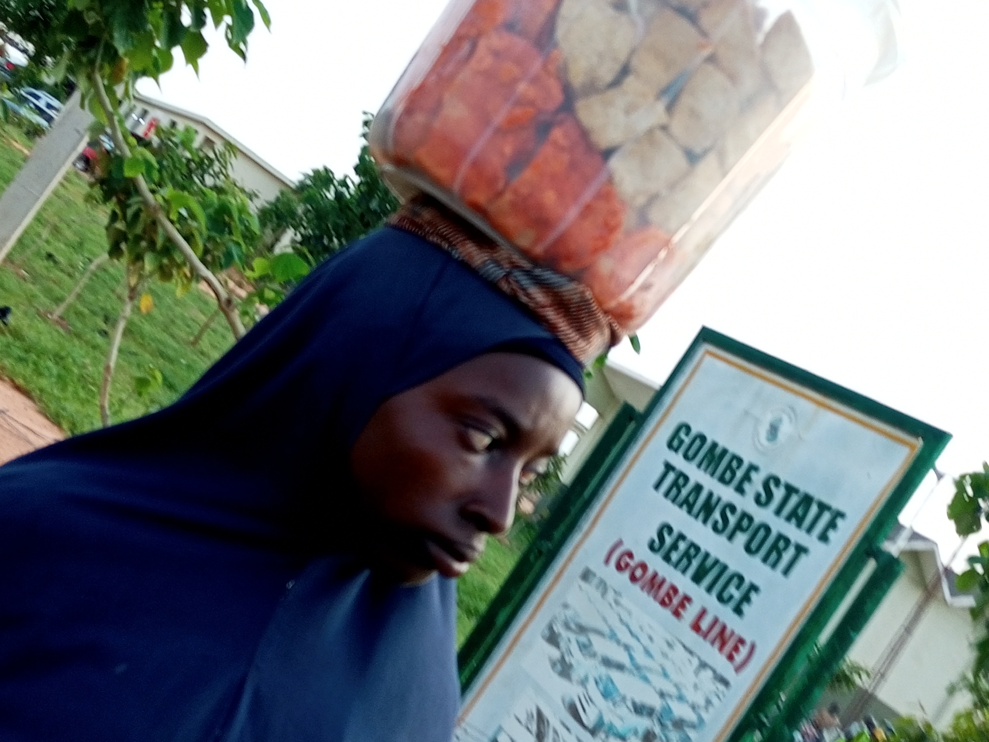 A close-up, upward angle shot shows a person wearing a blue hijab carrying a clear plastic container filled with food items on their head. The container is tilted, revealing a mixture of red and beige cubes, possibly food. The person's face, partially visible in profile, shows their eyes looking down and their mouth slightly open. A patterned scarf is wrapped around their forehead, beneath the container. In the background, a sign with green trim reads "GOMBE STATE TRANSPORT SERVICE (GOMBE LINE)" in green and red text. The surrounding environment includes greenery, trees, and what appears to be a building with white siding. The overall scene suggests a bustling urban environment in Gombe, Nigeria, likely a street or market area where people transport goods.