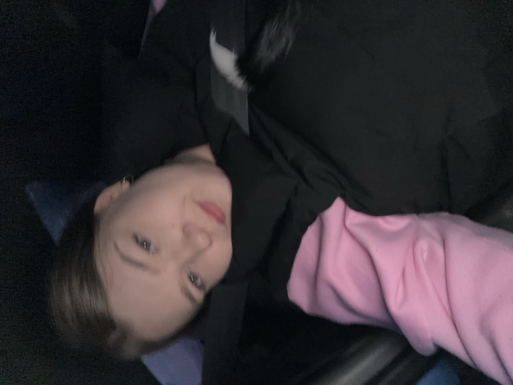 A young woman with fair skin, light eyes, and light brown hair is seated in a car. She wears a pink hooded sweatshirt under a black jacket with a fur trim and a seatbelt is across her torso. The interior of the car appears dark and out of focus. There is no indication of the location being Ольховка, Russia.