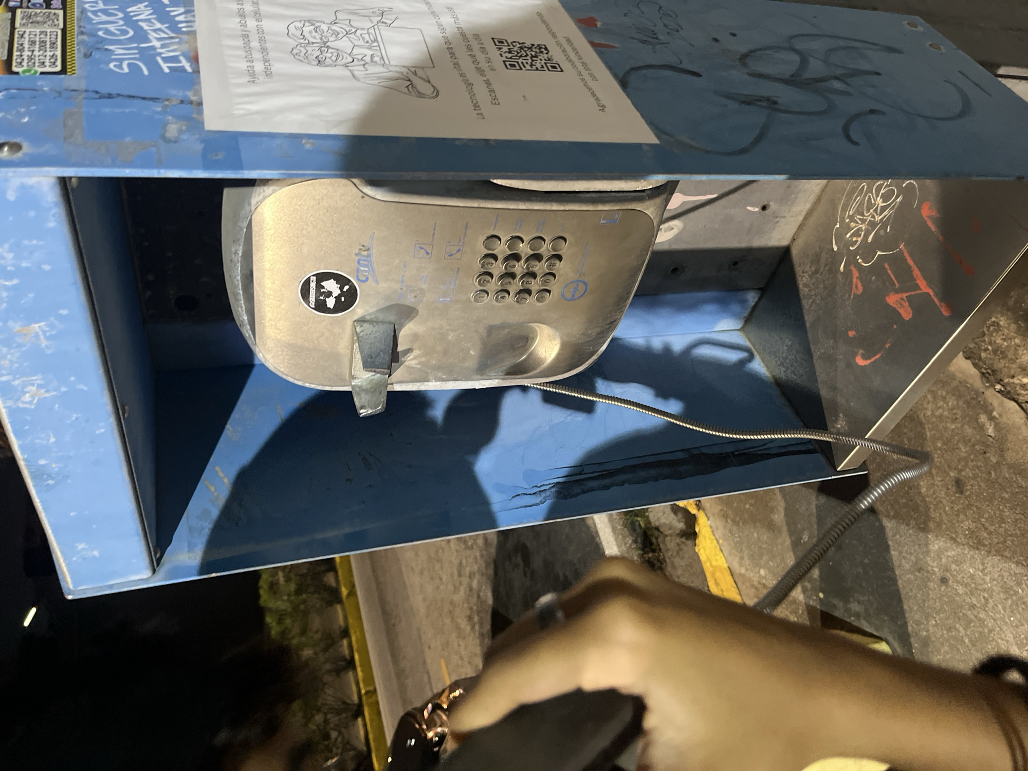 The image captures a public payphone booth in Pampatar, Venezuela. The booth itself is constructed from blue metal, showing signs of wear and graffiti. Inside, a silver payphone unit is mounted, featuring a keypad and a receiver. A coiled cord connects the receiver to the unit. A hand, possibly holding a mobile phone, is visible in the foreground, suggesting an interaction or comparison between the payphone and a modern communication device. The background shows a dimly lit street or parking area with some vegetation and a yellow curb. A white flyer with illustrations and text is affixed to the exterior of the booth. The overall scene depicts a juxtaposition of older and newer communication technologies in an urban setting.