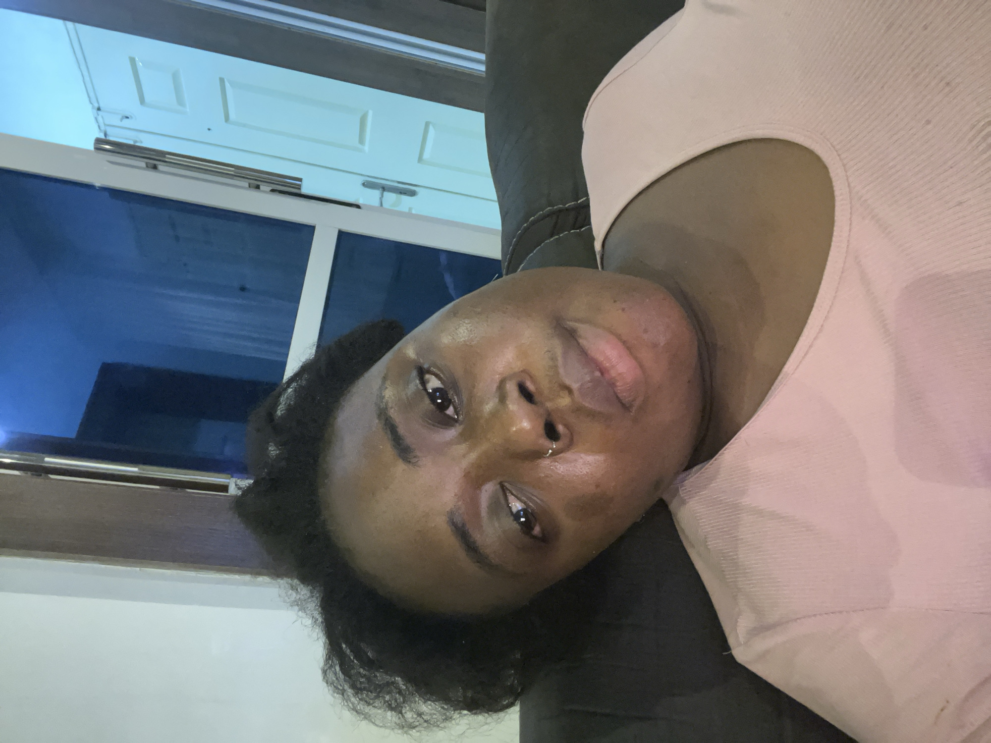A close-up, rotated portrait photograph shows a Black person with short, dark, curly hair. The person has brown eyes, a nose ring, and appears to be wearing a pink, ribbed shirt. The person is reclining on a dark-colored, possibly leather, couch. Behind the person is a window with white framing and a white door with a horizontal bar. The window is dark blue from the light source and reflection. The location and city of Jos, Nigeria cannot be confirmed.