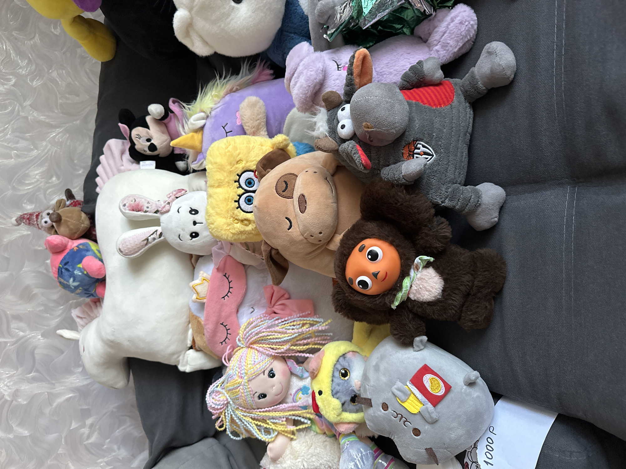 A collection of stuffed animals is arranged on a dark gray sofa, which appears to be indoors. The background is a white textured wallpaper. The stuffed animals are diverse in size, color, and character, including a large white unicorn, a plush dog, a purple unicorn, a yellow Spongebob Squarepants character, a Minnie Mouse plush, a brown bear, a pink sleep mask, a doll with colorful hair, a small grey animal wearing a yellow duck hat, a grey plush resembling a potato with fries, a small brown creature with large orange eyes, and a grey cow-like stuffed animal wearing a grey garment with red accents. A piece of white paper with "1000 P" written in black marker is placed on the sofa in the lower left corner of the image. The lighting suggests it is daytime. The location is stated to be the city of Chaikovsky, Russia.