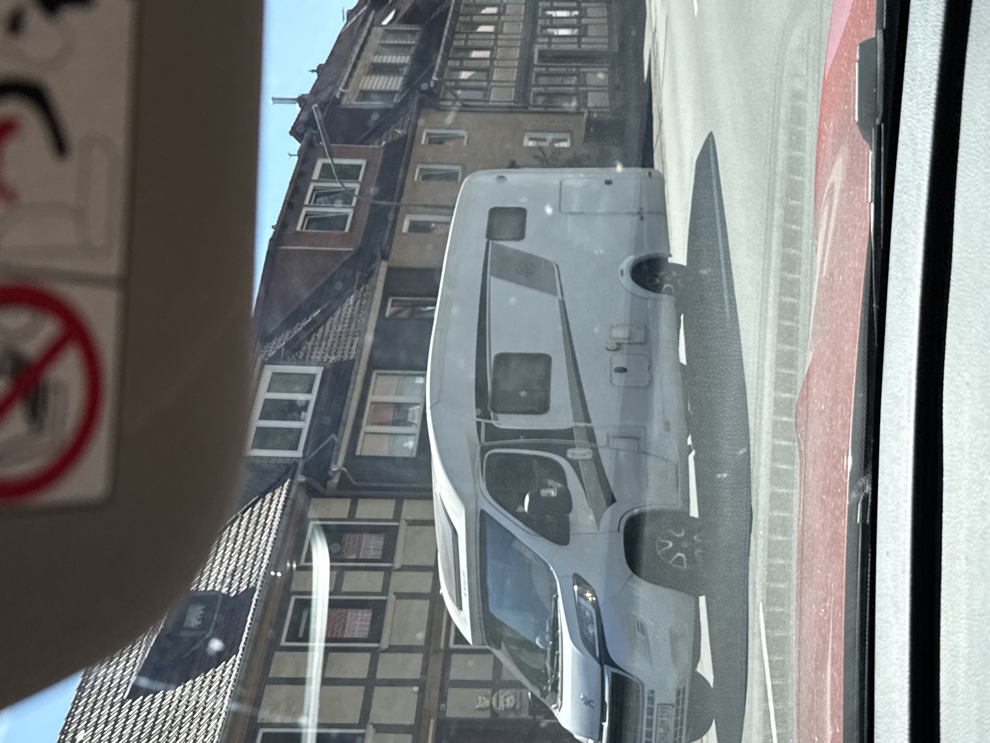The image shows a white campervan driving on a street in Bad Lauterberg im Harz, Germany. The campervan is in the foreground, and the background shows traditional half-timbered buildings. The sky is blue, and the weather appears to be sunny. The scene is captured from the perspective of someone inside another vehicle, looking through the windshield.