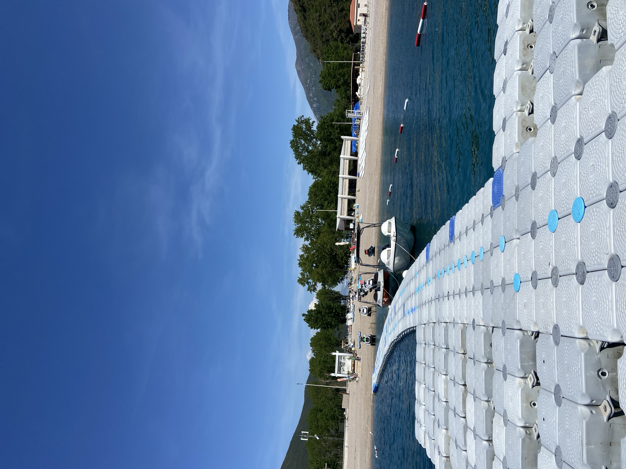 This image captures a serene, sunlit coastal scene in Prijevor, Montenegro, viewed from the water looking towards the shore. A prominent white modular floating dock, adorned with blue and grey circular accents on its surface, extends from the foreground into the midground on the right side of the frame. The dock appears to be largely empty of people.
To the left of the dock, calm, clear blue-green water reveals gentle ripples. Several red and white cylindrical buoys are scattered in the water, marking an area near the shore. A small, white boat is partially visible, seemingly docked alongside the floating platform.
The coastline features a pebbly or sandy beach, sparsely populated with a few indistinct figures. The beach is equipped with numerous white sun loungers and umbrellas, and several white, open-sided structures, possibly pergolas or beach kiosks, are visible. Further inland, a lush expanse of green trees lines the shore, and in the background, a series of towering, forested mountains rise dramatically under a vast, cloudless blue sky, save for a few faint wisps of clouds in the upper left. A building with a reddish roof can be seen amidst the trees on the far right of the beach, and another white structure with a blue slide is visible further down the coast. The scene suggests a beautiful, peaceful day at a coastal resort. No visible text is discernible.