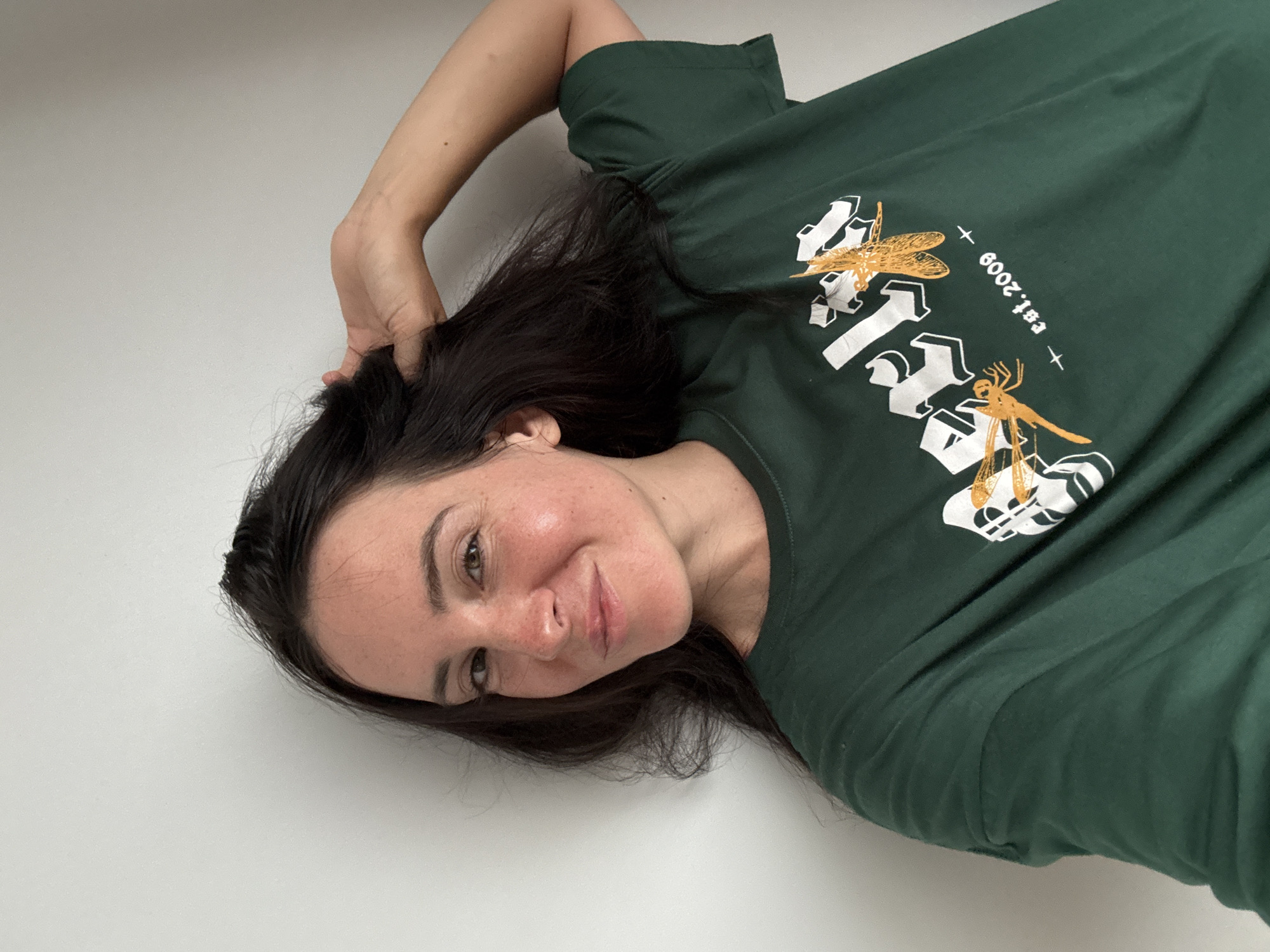A woman with long, dark hair and fair skin is pictured in a medium shot, looking at the camera with a slight smile. She is wearing a dark green t-shirt with a graphic design featuring dragonflies and the text "est. 2009". Her right hand is raised and playing with her hair. The background is a plain, light-colored wall. The image does not provide any information about a location in North Kuta, Indonesia.