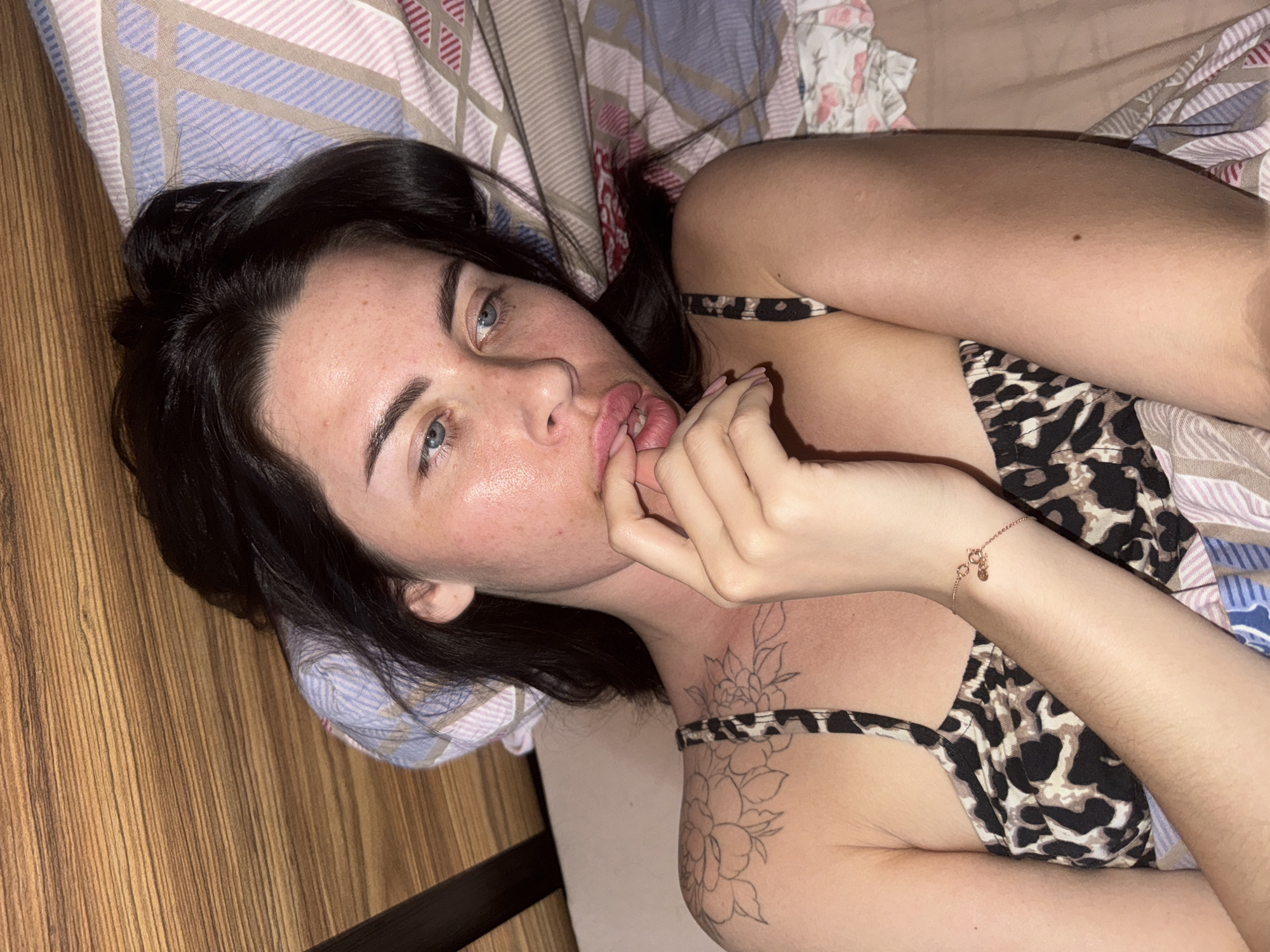 A woman with dark hair and blue eyes is lying on a bed with patterned bedding. She is wearing a leopard print top and has a tattoo of flowers on her left shoulder. She is holding her hand up to her mouth. The room appears to be dimly lit. The image does not provide any information to suggest a location in Chaikovsky, Russia.