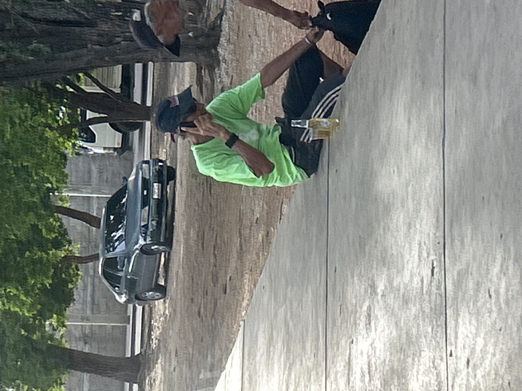 The image depicts an outdoor scene, likely in Santa Ana, Venezuela, featuring a man seated on a concrete ledge. The man, wearing a bright green t-shirt and black shorts with white stripes, is holding a mobile phone to his ear. Next to him, on the ledge, are two bottles of beer and a glass, one bottle partially full. In the background, a dark-colored sedan is parked, and further behind it, a white truck is visible. The environment includes trees with lush green foliage and a dirt or gravel ground surface. Another person, mostly obscured from view and wearing a dark hat, is partially visible in the right side of the frame, interacting with a dark-colored object, possibly an animal, which is also partially visible. The lighting suggests it is daytime, with some shadows indicating sunlight.