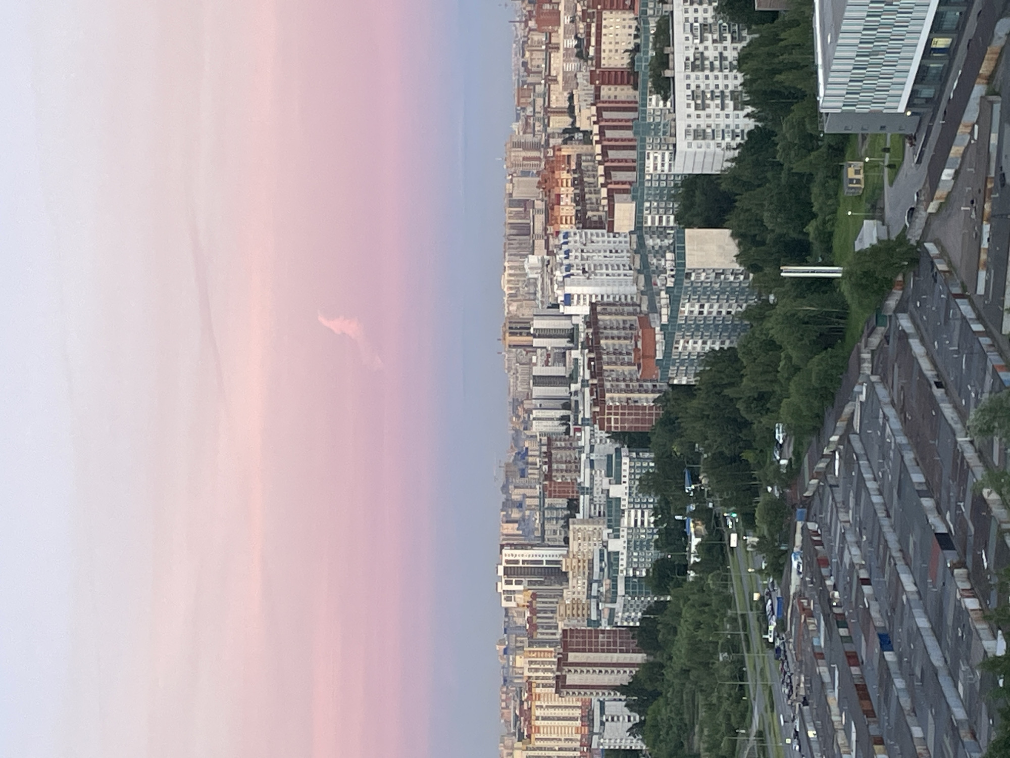 The image shows an aerial view of a dense urban landscape at dusk. The sky is a soft gradient of pink and purple, with a wispy cloud formation near the center. Below the sky, a sprawling city is visible, characterized by numerous multi-story buildings of varying architectural styles and colors. The buildings are packed closely together, creating a textured urban fabric. In the foreground, there is a large area filled with rows of individual garages, most of which appear to be made of corrugated metal and are weathered. Trees and green spaces are interspersed throughout the city, particularly in the middle ground, contrasting with the built environment. A road with several cars is visible between the garages and the residential buildings. The overall impression is of a large, residential city at the end of the day.
