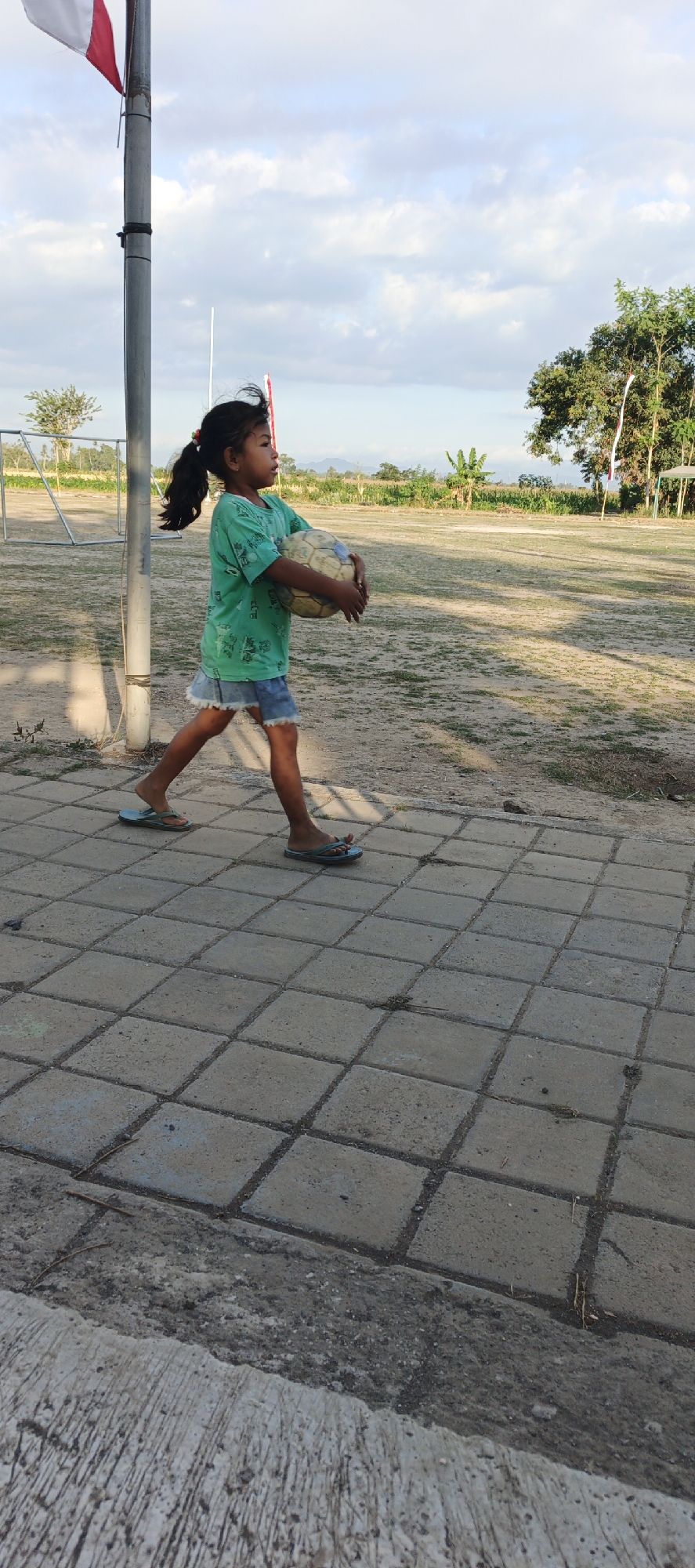 A young Indonesian girl walks along a paved walkway, holding a soccer ball. She is wearing a green t-shirt and a denim skirt. The walkway is made of square concrete tiles and is adjacent to a dirt area. In the background, there is a flagpole with a red and white flag, a soccer goal, trees, and fields of crops. The sky is cloudy. The scene appears to be in Kasiyan Timur, Indonesia.