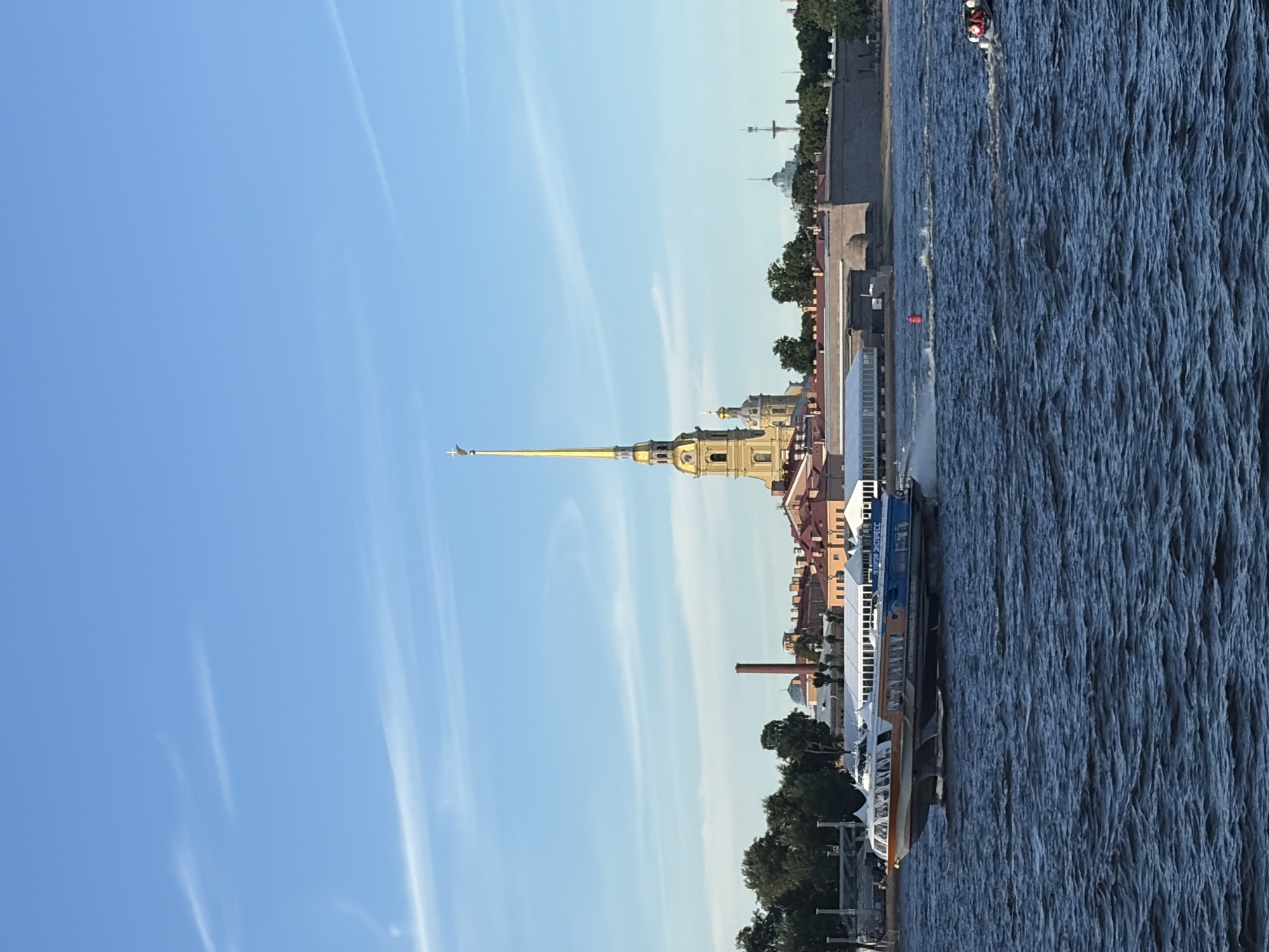 This is a full-shot, eye-level, natural-light photograph taken on a sunny day overlooking the Neva River in Saint Petersburg, Russia. In the foreground, the choppy, deep blue water of the river dominates the lower half of the frame. A hydrofoil passenger boat, the "Petrop Express," with an orange and blue color scheme, speeds across the water from left to right, leaving a white wake. In the midground, the Peter and Paul Fortress complex is visible on the far bank. Its prominent golden spire, topped with a weather vane, rises high above other buildings, including a yellow bell tower and several red-roofed structures. A red buoy is visible in the water between the hydrofoil and the fortress. Further in the distance, behind the fortress, are trees and other buildings, including a brick chimney and domes. The sky above is a clear, bright blue with scattered wispy white clouds. To the far right, a small boat with several occupants is moving away from the viewer.