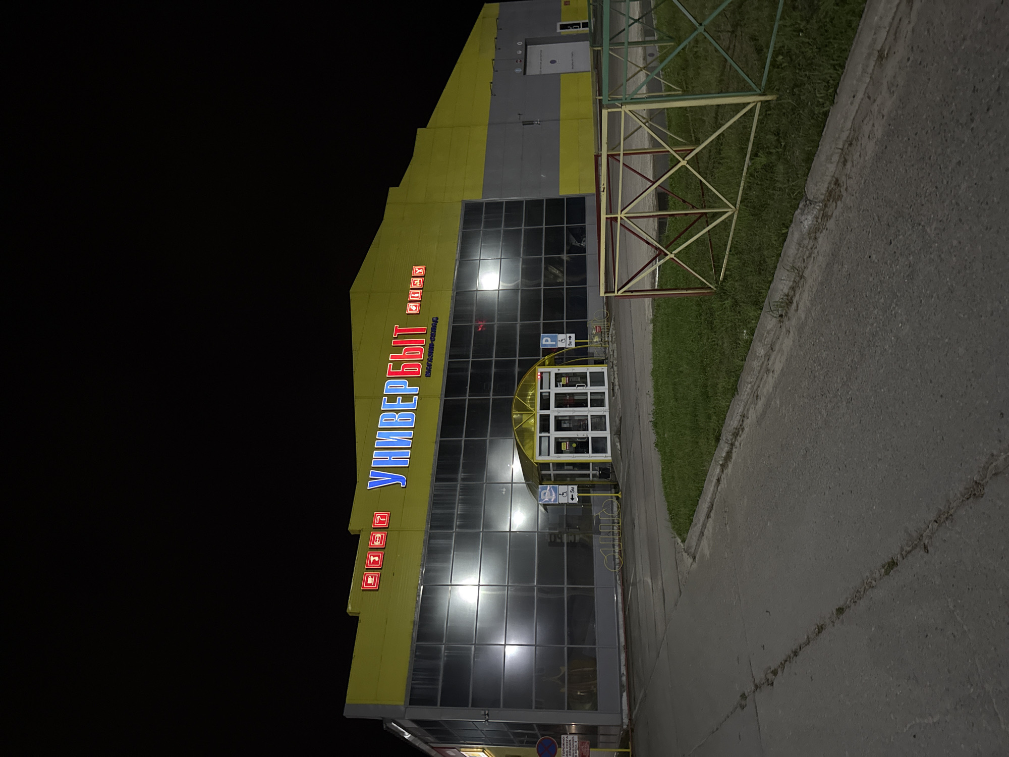 The image shows a nighttime exterior view of a large, modern building with a bright yellow and grey facade. The building is identified by large, illuminated blue and red text that reads "УНИВЕРБЫТ" (Univerbyt), likely the name of a department store or supermarket, with smaller red icons above the text. The building features a large, glass-paneled section reflecting interior lights. An entrance area with a yellow awning and glass doors is visible, with signs indicating parking and accessibility. A grassy area with a decorative metal fence runs along the front of the building. The foreground shows a cracked asphalt surface, possibly a parking lot or walkway, with some white markings on the ground. Streetlights or the building's own lighting illuminate the scene. The location is identified as Sovetskiy, Russia.