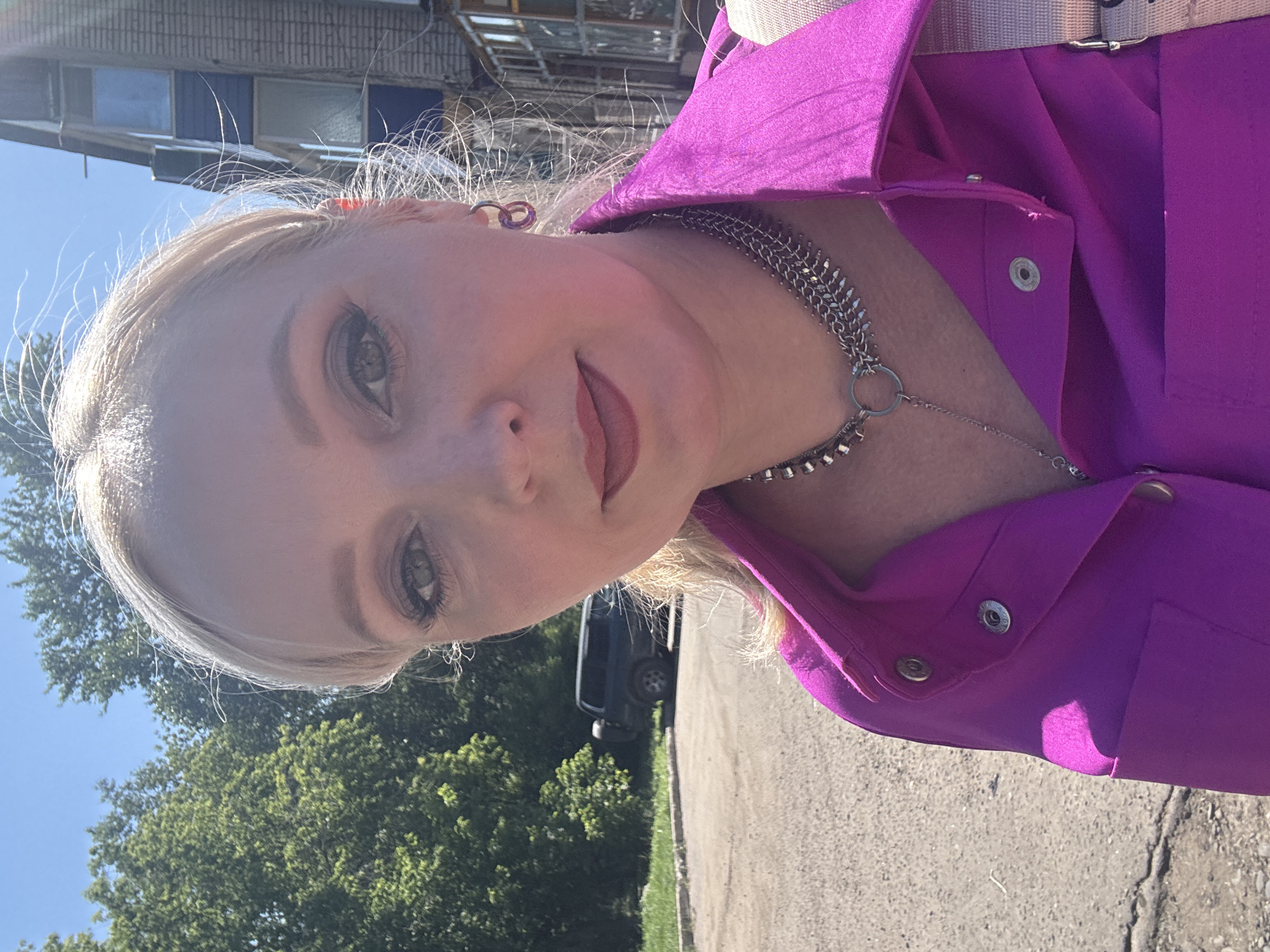 The image is a close-up, selfie-style shot of a blonde woman in Komsomolsk-on-Amur, Russia, taken outdoors during the day. She is wearing a vibrant fuchsia shirt with a collar and snap closures, layered with a multi-strand silver necklace featuring a circular pendant. Her hair is blonde and styled with some wisps blowing in the wind. She is wearing makeup, including eyeshadow and lipstick. In the background, a dark-colored SUV is parked on a gravelly area adjacent to a tree-lined street. Behind the trees, a multi-story brick building with windows is visible. The sky is clear and blue. The overall setting appears to be a residential or commercial area within the city.