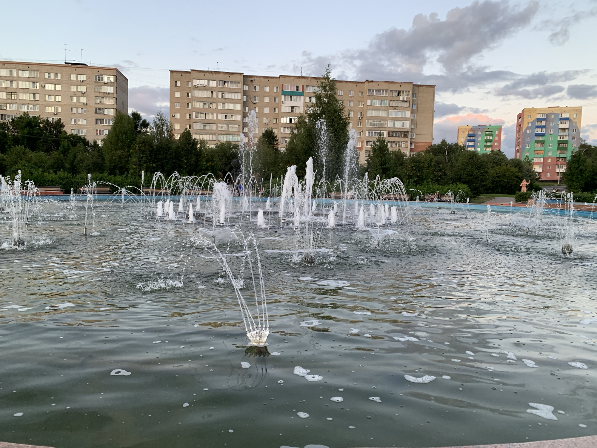 The image shows a public fountain in the city of Prokopyevsk, Russia, with apartment buildings in the background. The fountain is active, with water jets spraying upwards, creating a dynamic scene. The water in the fountain is dark green and appears somewhat murky, with some foam on the surface. In the background, several tall, multi-story apartment buildings are visible. The buildings vary in color and architectural style, with one appearing to be a light beige color and another featuring a colorful, geometric facade in shades of blue, green, yellow, and pink. Trees and greenery are interspersed between the buildings and surrounding the fountain area, suggesting a park-like setting within the urban environment. The sky is overcast with soft, diffused light, indicating it might be late afternoon or early evening. There are no people clearly visible or engaged in any significant activity in the foreground or mid-ground of the image. The overall scene depicts a common urban public space designed for recreation and aesthetics.