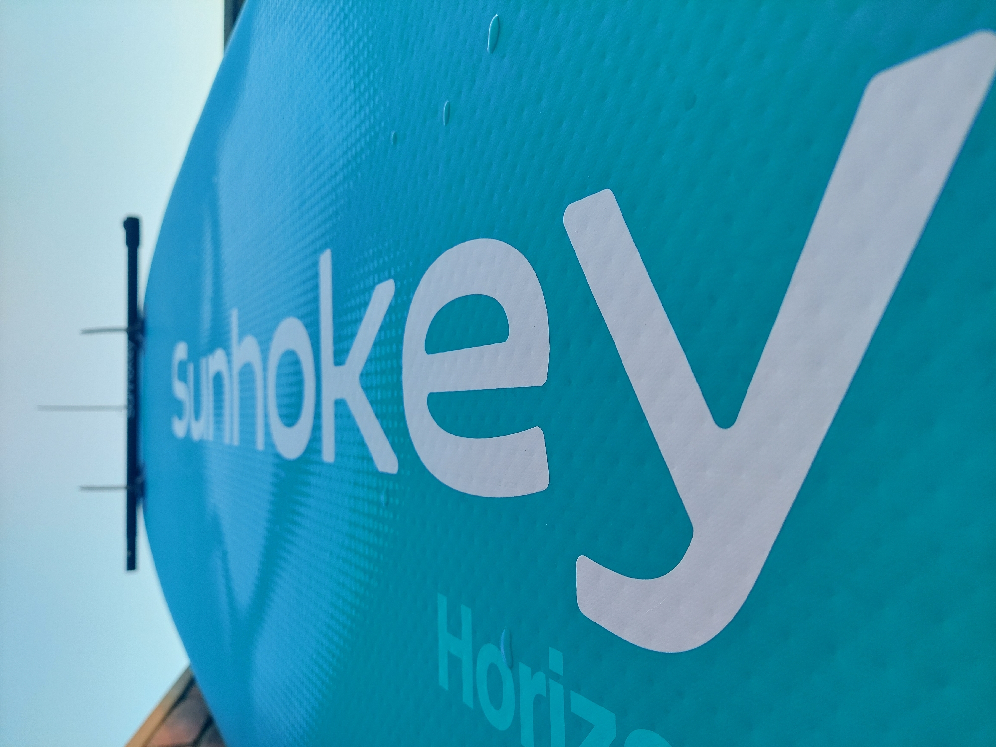 The image is a low-angle, close-up shot of a teal blue paddleboard. Large white lettering on the paddleboard reads "re smoke" with a partial "Horiz" visible on the left side. The surface of the paddleboard has a textured grip pattern and a few small water droplets. In the background, the sky is a clear pale blue, and a portion of a brick structure is visible on the left. The paddleboard appears to be resting on a surface outdoors, possibly near the water, given its nature as a paddleboard. The specific location in Pirallahi Raion, Azerbaijan, cannot be determined from the visual information present in the image. There are no people or animals visible.