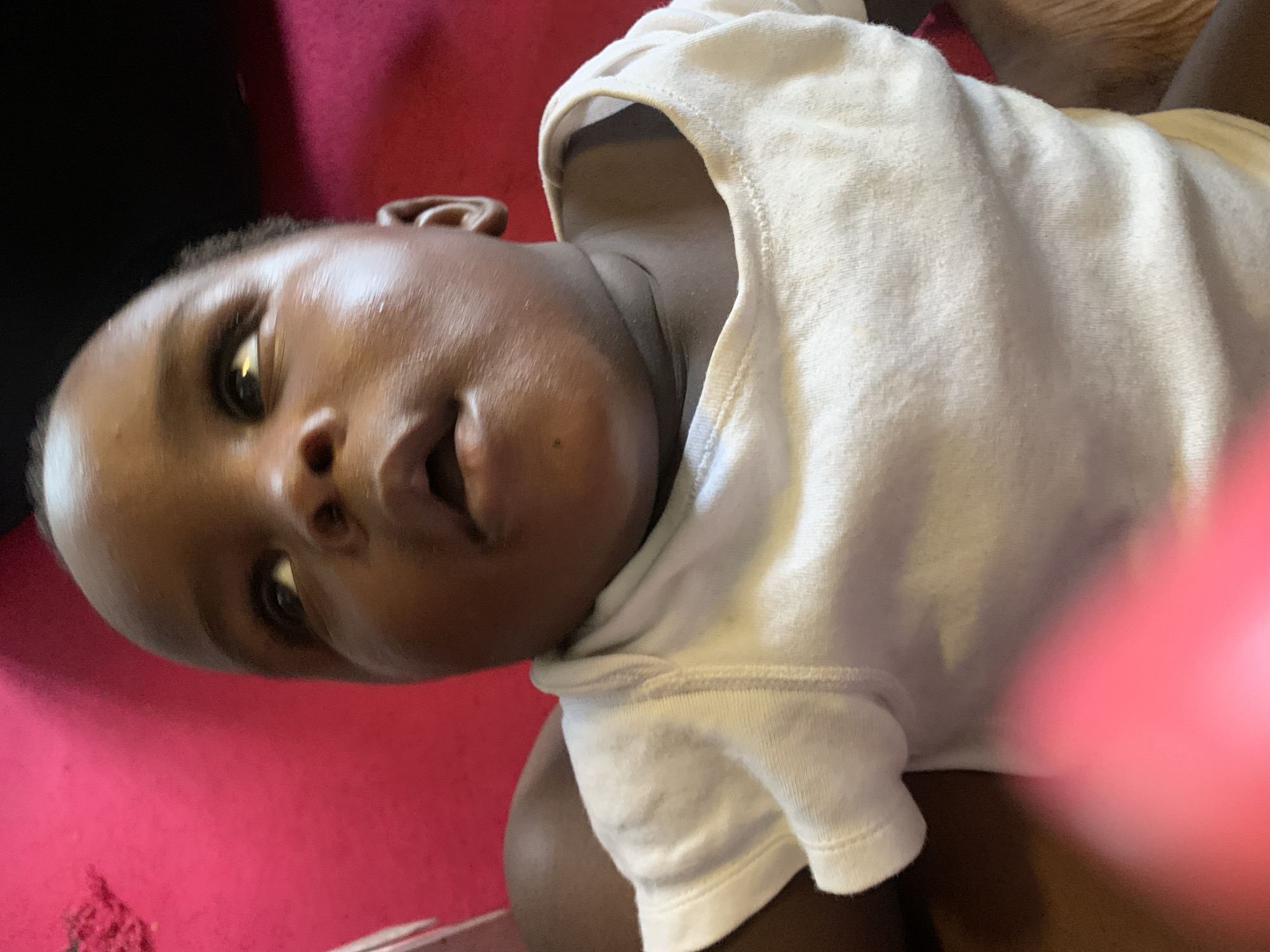A close-up shot focuses on the face and upper body of a Black infant, who is looking upwards with their mouth slightly open. The infant is wearing a white short-sleeved onesie, and their skin appears smooth and rosy, with a slight sheen. The background is a vibrant, textured pink surface, possibly a blanket or cushion. To the right of the infant, a darker, blurred area suggests another surface or person. A bright pink, out-of-focus object is visible in the lower left corner of the frame, partially obscuring the infant's chest. The lighting is bright and casts soft shadows on the infant's face, highlighting their features. The image is taken from a slightly low angle, looking up at the infant.