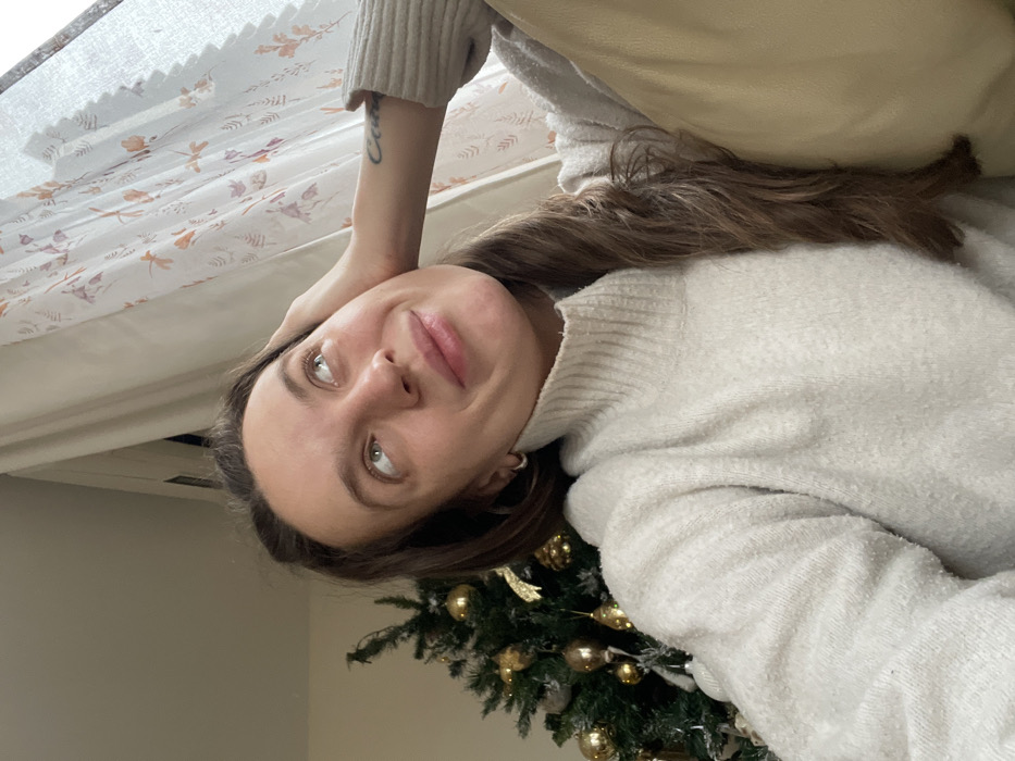 The image displays an adult female indoors, captured in a selfie-style orientation. She possesses long, wavy, dark brown hair and a light complexion, looking upwards and to the right with a subtle smile. Her left hand rests on the side of her head, above her ear, with a dark text tattoo visible on her left forearm. She is dressed in a light-colored, textured turtleneck sweater and small silver hoop earrings. In the background to her left, a portion of an artificial Christmas tree is visible, adorned with multiple gold spherical ornaments and gold tinsel. To her right, a window is covered by light-colored curtains patterned with leaves and orange insect-like figures. A light beige upholstered cushion is present in the lower right foreground. The specific location within Budva, Montenegro, cannot be determined from the visual evidence provided in the image.