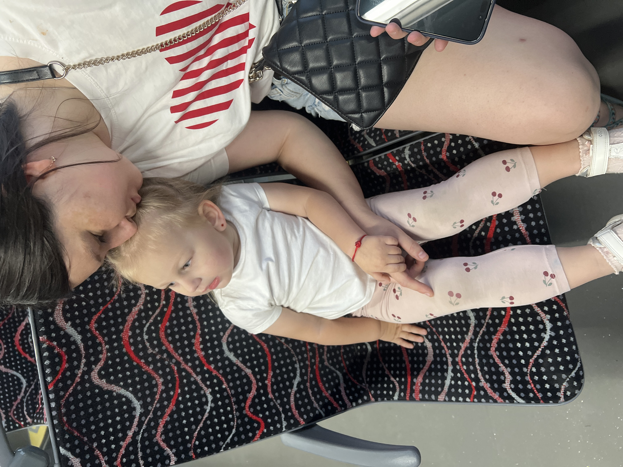 In Pilsen, Czechia, a woman is seated next to a young child on a patterned seat. The woman is leaning down, touching the child's forehead with her own, and appears to be kissing the child. The child, with blond hair, is wearing a white t-shirt and light pink leggings with a cherry pattern. A red string bracelet is visible on the child's wrist. The woman is wearing a white t-shirt with a red and white striped heart design and denim shorts. She has a black quilted handbag on her lap and is holding a smartphone. The background shows the patterned upholstery of the seat.