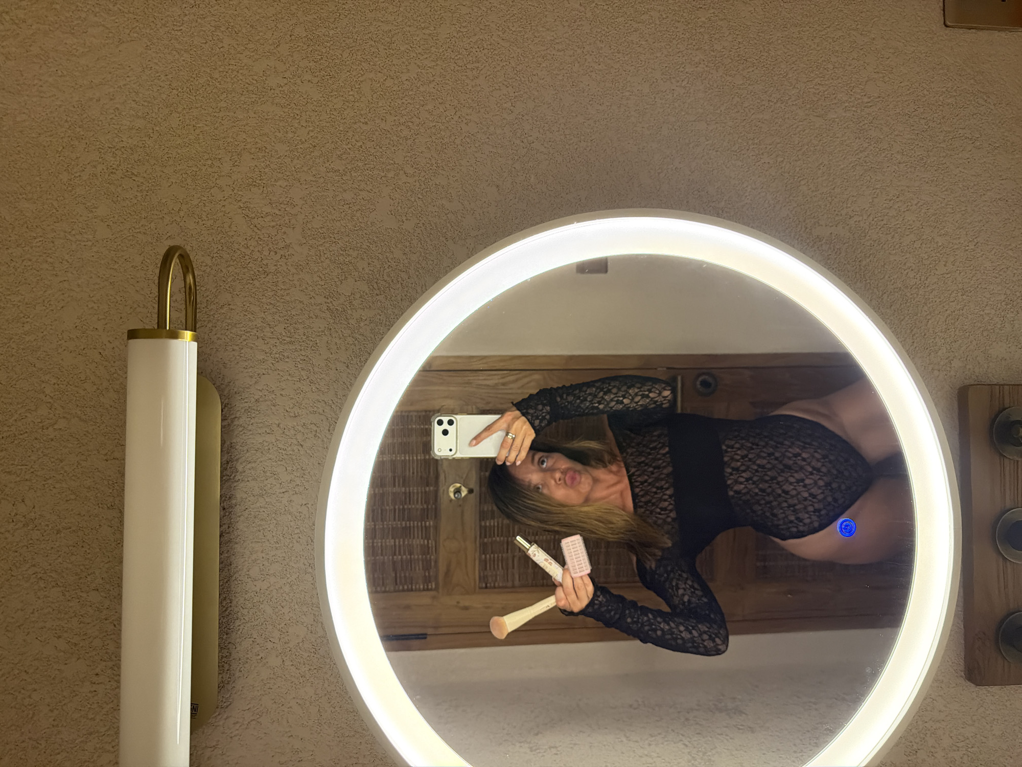 A woman is taking a mirror selfie in a bathroom. She is wearing a black lace bodysuit and holding makeup brushes and a bottle of what appears to be a beauty product. The mirror is circular and illuminated by a bright LED light. Behind her, a wooden door is visible. The wall has a textured finish, and a light fixture is mounted above the mirror. The image does not contain enough information to determine the specific location in Mengwi, Indonesia.
