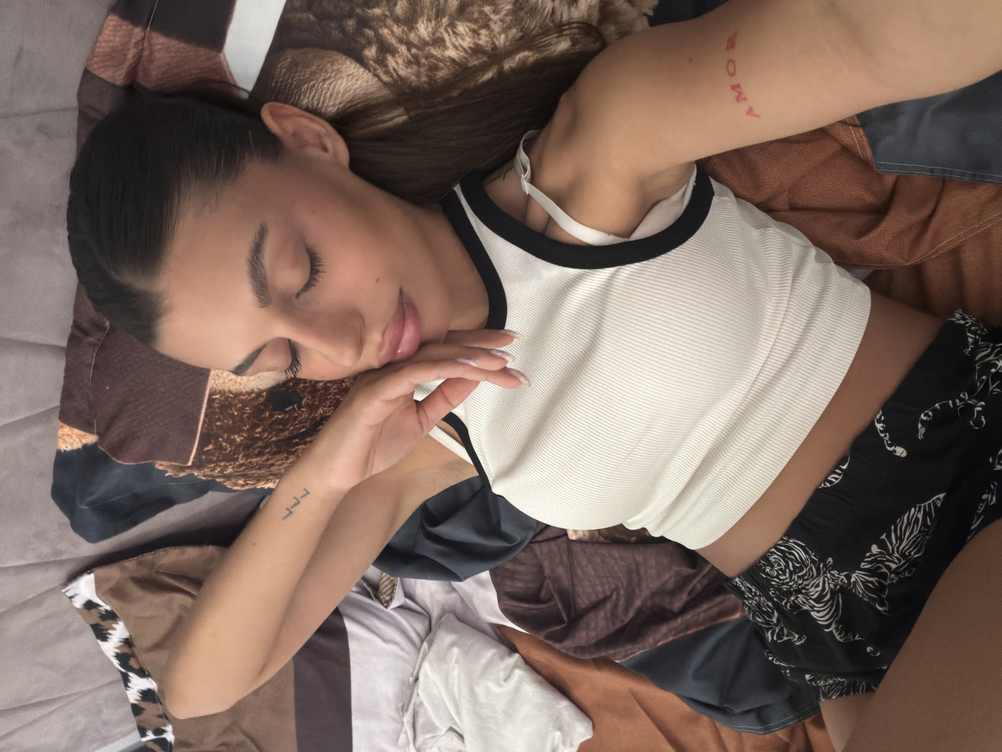 The image shows a close-up, overhead view of a young woman lying on a bed. She has her eyes closed and is resting her head on her hand, with her face angled slightly to the right. She has dark hair pulled back into a ponytail and is wearing a white ribbed tank top with black trim and black shorts with a white tiger pattern. There are two tattoos visible on her arms: a small "LL L" on her left wrist and the word "AMOR" in red on her right bicep. The bed is covered with patterned bedding in shades of brown, beige, and black, and some white and grey fabric is visible in the lower left corner. The scene is indoors, likely a bedroom, but no specific city or location is discernible from the image itself.