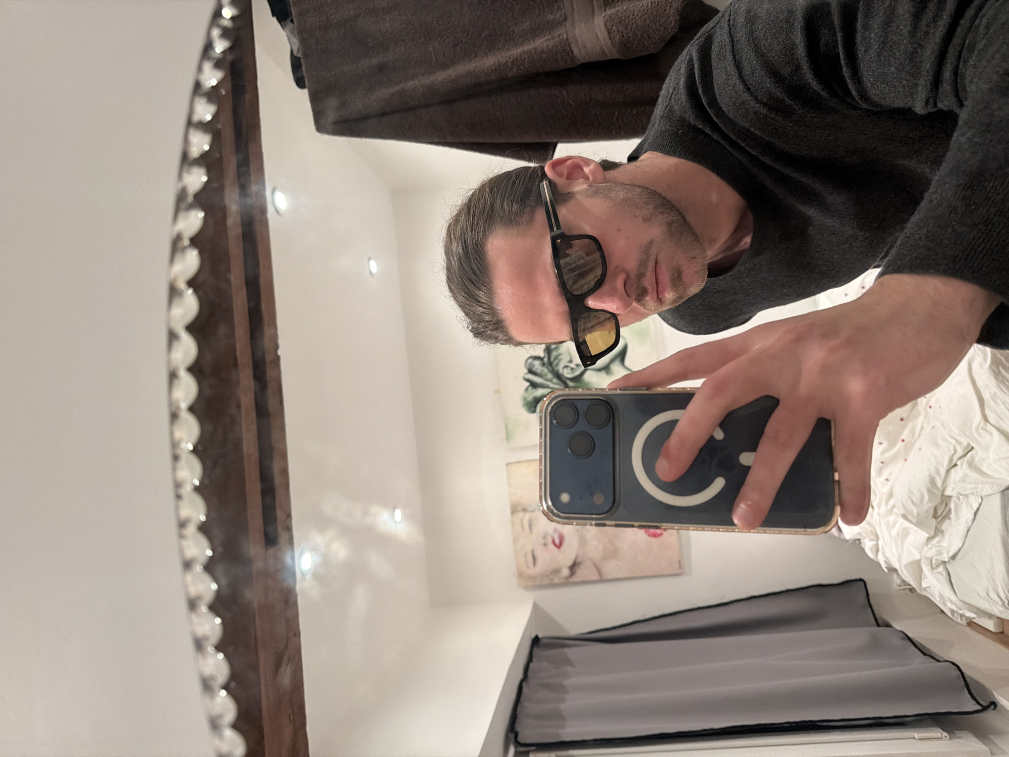 A male subject is captured in a mirror selfie within a residential interior in Paris, France. The subject, seen from the upper torso, wears a dark long-sleeved sweater and black sunglasses with yellow-tinted lenses. His dark hair is pulled back, and he has a trimmed beard. His right hand holds a dark-colored iPhone, fitted with a clear case featuring a white MagSafe ring, oriented to photograph his reflection. The mirror has a decorative, scalloped top edge. The reflected background reveals a white wall adorned with two artworks: a pop-art style portrait of a woman with blonde hair and red lips, and a subsequent green-toned abstract piece. Above, a white ceiling contains recessed spotlights and a visible wooden structural beam. To the left, a grey curtain with black trim partially covers a window or opening, below which a white radiator is present. A bed with crumpled white bedding, featuring small red patterns, is visible in the lower left. A dark brown towel is draped over an unseen object to the subject's right. The architectural features, including the wooden beam and radiator, are consistent with an older Parisian apartment setting.
