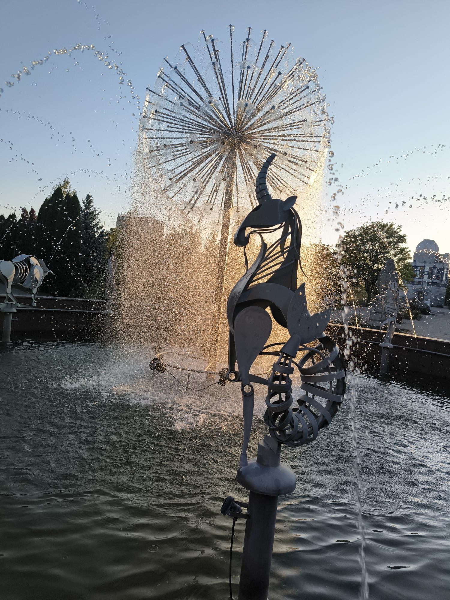 The image depicts a fountain with a distinctive dandelion-like sculpture at its center, from which water sprays outward. In the foreground, a dark metal sculpture resembling a stylized horse or unicorn is positioned in front of the main fountain element. The water is illuminated by the warm glow of the setting sun, creating a golden hue and numerous water droplets suspended in the air. The fountain is situated in a plaza with paving stones visible in the background, along with trees and buildings. To the left, another metallic sculpture resembling an animal, possibly a bull or ox, can be seen. The overall scene suggests an outdoor public space, likely a park or square, in Chișinău, Moldova, given the context of the prompt. The sky is clear and blue, indicating good weather. There are no people visible in the image.