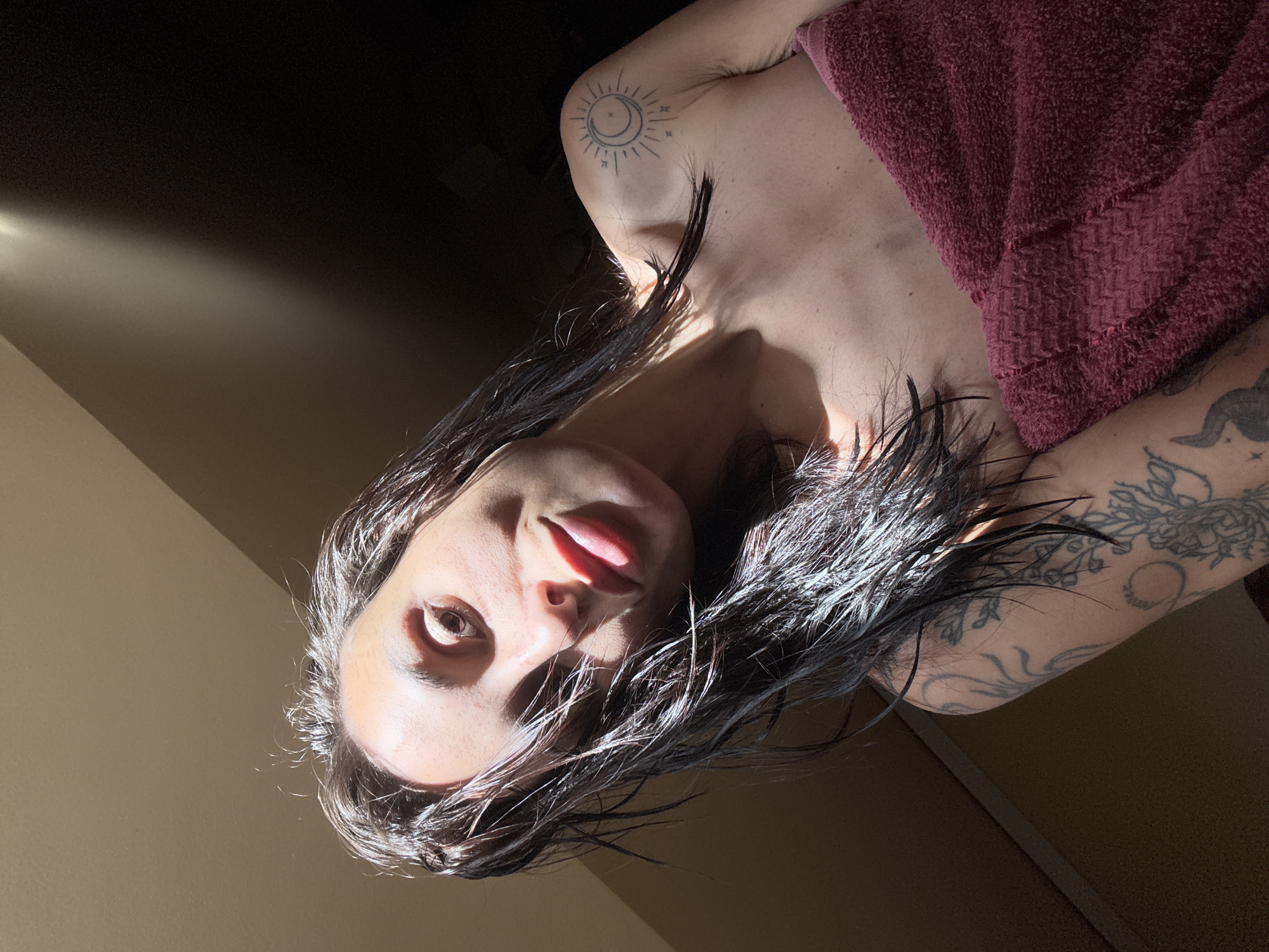 A person with wet, dark hair is positioned in a dimly lit room. The person is facing the camera with their tongue slightly sticking out. A maroon-colored towel is draped over one shoulder. The person has tattoos on their upper arm and shoulder. The location is unknown and there is no indication of Pleven, Bulgaria.