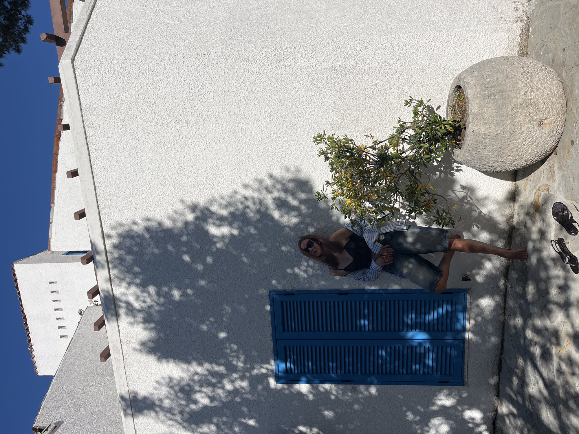 The image displays an outdoor scene in Budva, Montenegro, under bright sunlight, with the composition notably rotated 90 degrees clockwise from its likely original orientation. For clarity, the description assumes a standard vertical orientation.
A woman is depicted resting against a white, textured stucco wall. She has long, light brown hair and wears dark sunglasses, a black top, denim shorts, and an open, light-colored striped button-down shirt. Her bare feet extend onto the paved ground, where a pair of black sandals and what appears to be another pair of sunglasses are placed. She looks towards the viewer with a relaxed expression.
Below her, a prominent feature on the white wall is a vibrant blue window with closed louvered shutters. To the right of the woman, a large, round, grey stone planter containing a small, leafy green plant is situated on the paved ground. The ground itself is made of light-colored, possibly stone, paving.
The scene is characterized by strong, dappled shadows cast by unseen trees or foliage, falling across the white wall and the ground, indicating intense, clear sunlight, likely during midday. In the background to the left, parts of other white buildings with distinctive wooden beams and small rectangular openings are visible against a clear, deep blue sky. The architecture, colors, and lighting collectively evoke a quintessential Mediterranean coastal setting. No visible text is present in the image.