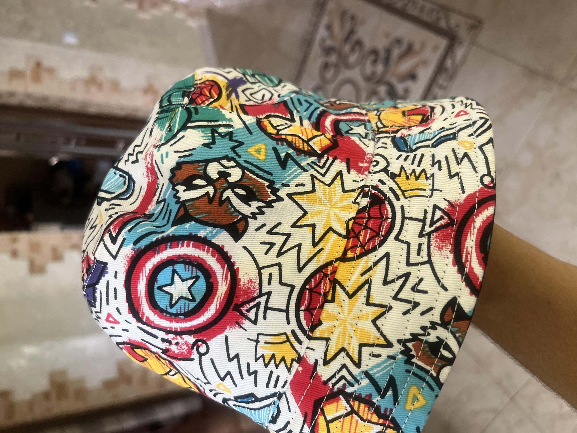 In Onokivtsi, Ukraine, a close-up, over-the-shoulder shot captures a person's hand holding a brightly colored bucket hat. The hat is adorned with a chaotic, comic-book-style pattern featuring superhero emblems like Captain America's shield, bursting stars, and abstract graphic elements in red, blue, yellow, and green. The background is softly blurred, showing a hint of a tiled floor and a muted, textured wall, suggesting an indoor domestic setting.