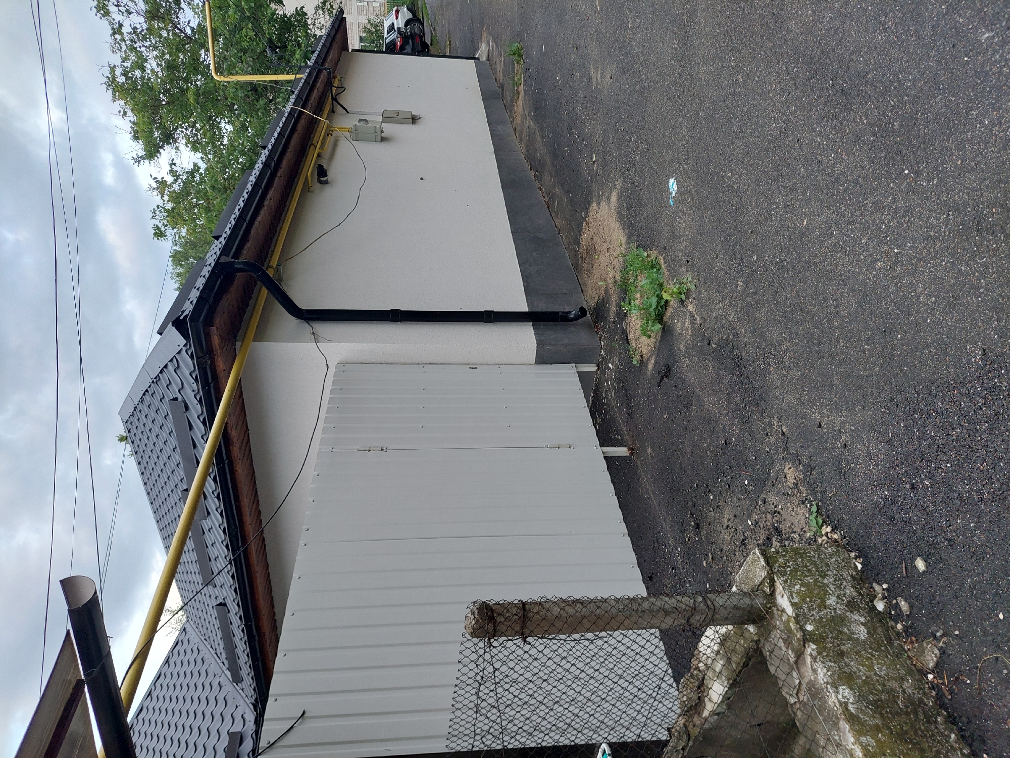 This image captures an exterior scene in Orhei, Moldova, under an overcast sky. The primary subject is the side of a single-story building featuring light-colored (off-white) textured stucco walls. A dark grey corrugated metal roof is visible, equipped with a black half-round gutter system and a black vertical downspout. Beneath the roofline, a light brown wooden soffit/fascia is present. The lower section of the building wall, approximately 0.5 meters high, is painted a darker grey.
Affixed to the building wall are utility elements, including a yellow gas pipe running horizontally below the roofline and ascending on the right. Two utility boxes, one off-white and one darker grey, are mounted on the wall, along with several black electrical wires and a small dark security camera.
To the left of the building, a white corrugated metal barrier or fence with vertical panels extends into the foreground, partially obscuring the structure. In the extreme foreground on the left, a segment of a chain-link fence is visible, attached to a moss-covered concrete post and a low, moss-covered concrete barrier.
The ground is covered with dark asphalt pavement, exhibiting both worn areas and segments that appear more recently laid. Small patches of green weeds and grass protrude through the asphalt, notably near the base of the downspout where a sandy patch is also visible. A small piece of light-colored litter lies on the asphalt in the mid-foreground.
In the background, green deciduous trees are prominent behind the building, and faint outlines of other structures and parked vehicles (including a white SUV and a dark car) are discernible on the far right. No individuals or animals are present in the scene.