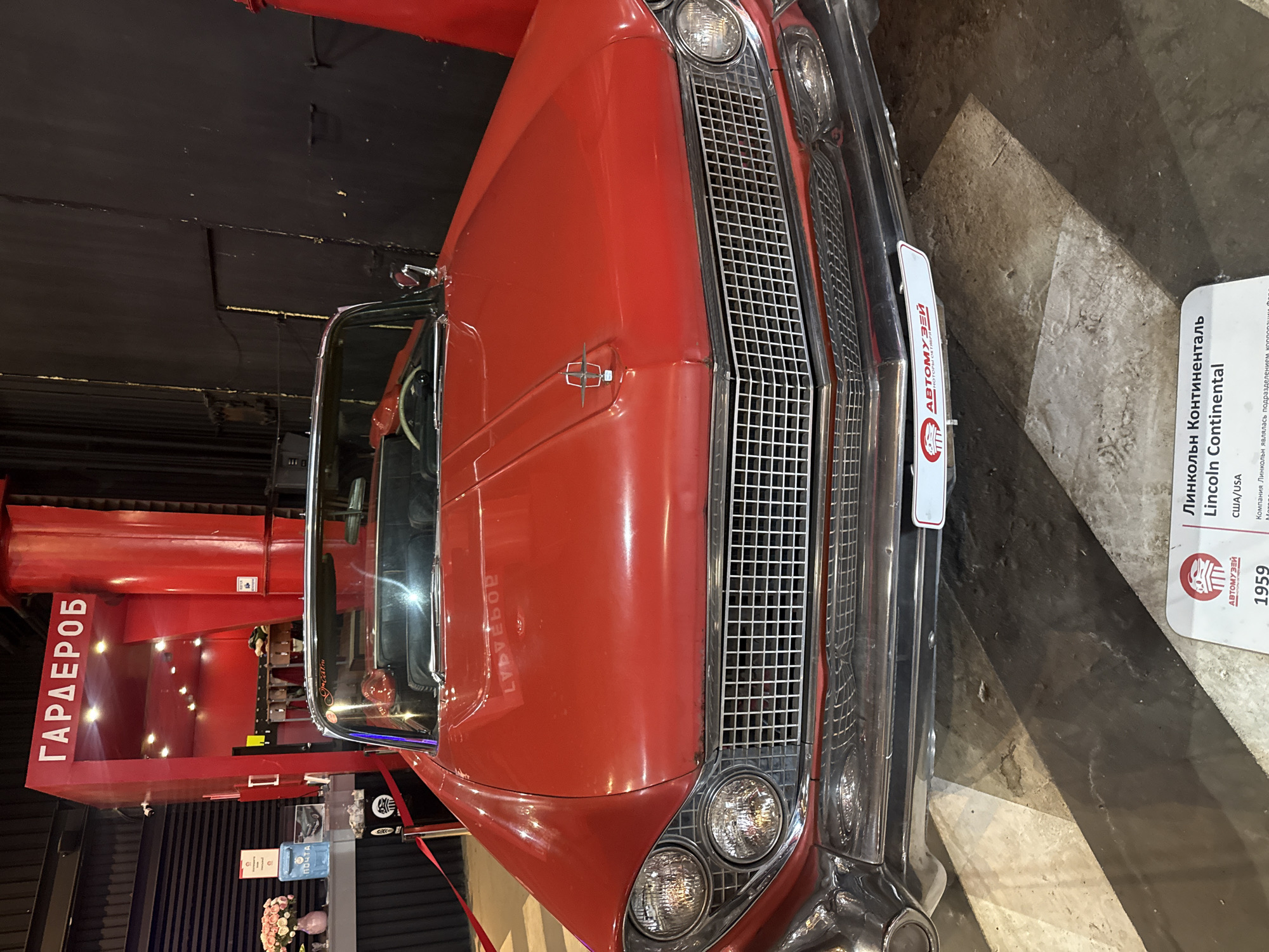 The image shows the front of a red 1959 Lincoln Continental convertible, parked indoors. Behind the car, a red structure with the Russian word "ГАPДЕРОБ" (Garderob, meaning cloakroom or wardrobe) is visible. This structure appears to be part of an exhibition or museum setting. To the left of the car, a blue mailbox with the word "ПОЧТА" (Pochta, meaning post office) and a vase of pink flowers are on display. The floor has a black and white striped pattern. A sign at the bottom of the image, partially obscured, identifies the car as a Lincoln Continental from the USA. The setting is likely a museum in Moscow, Russia, possibly related to automobiles or retro culture.